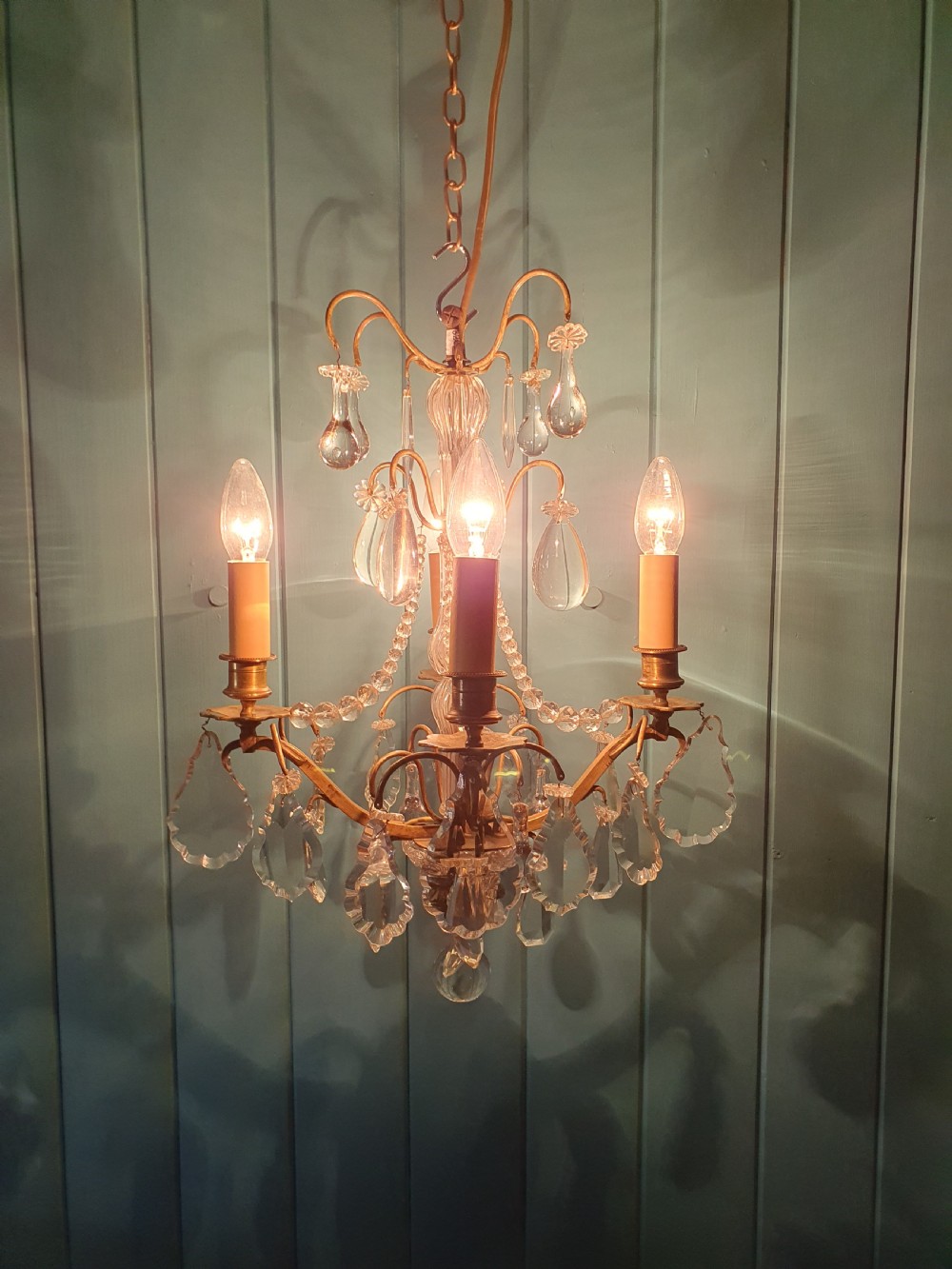 An antique French brass and crystal chandelier featuring curved scroll arms, a central glass stem, candle sleeves, and hanging teardrop glass prisms. View VI