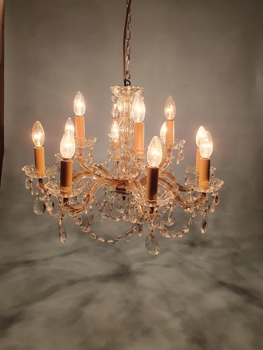 An early 20th-century two-tier crystal chandelier featuring twelve brass arms, glass drops, faux candle sleeves, and dark fabric shades. View V