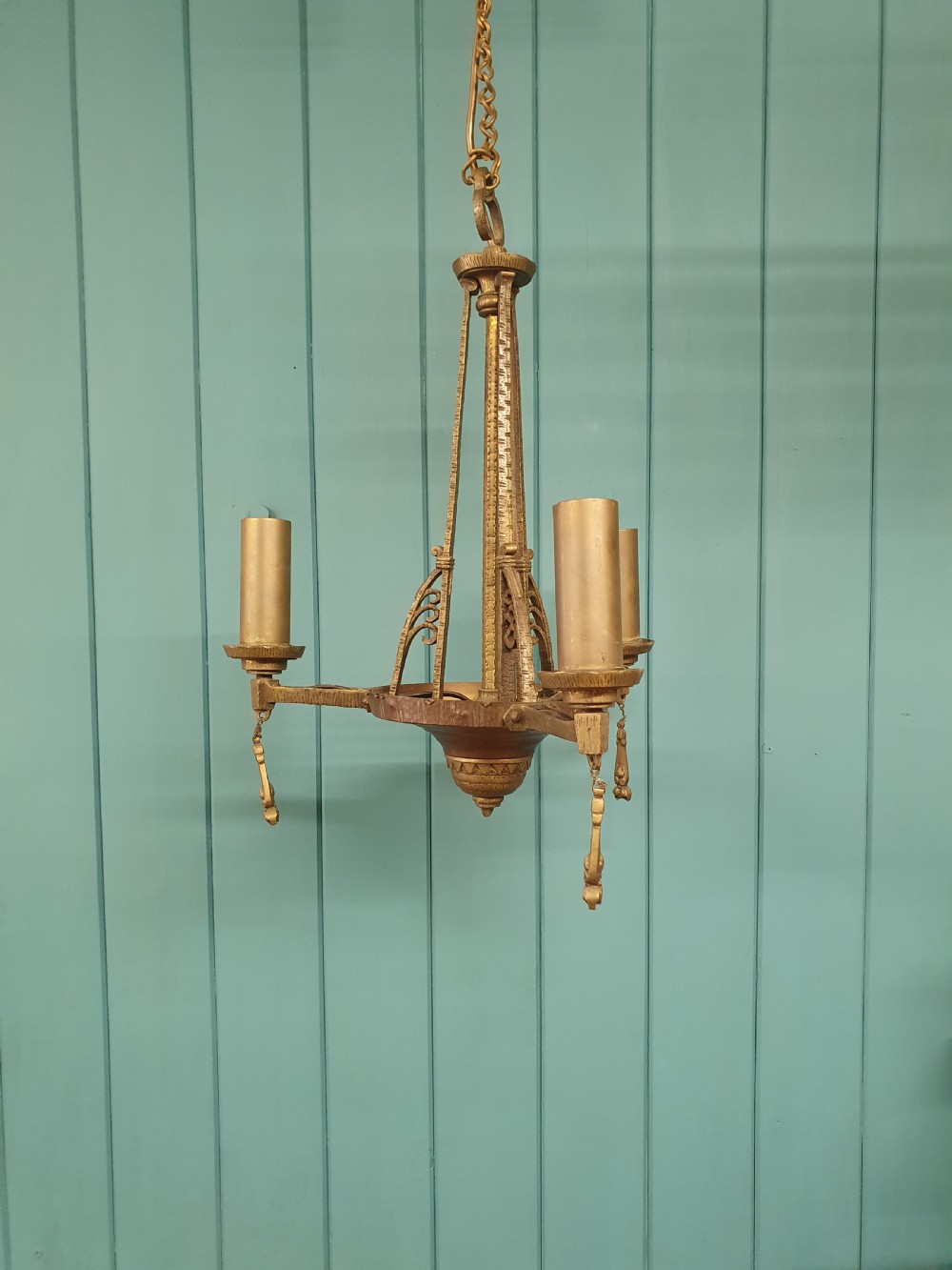 An Art Deco gilt brass three-arm chandelier featuring geometric central columns, candle-style bulb holders, and dangling teardrop accents.