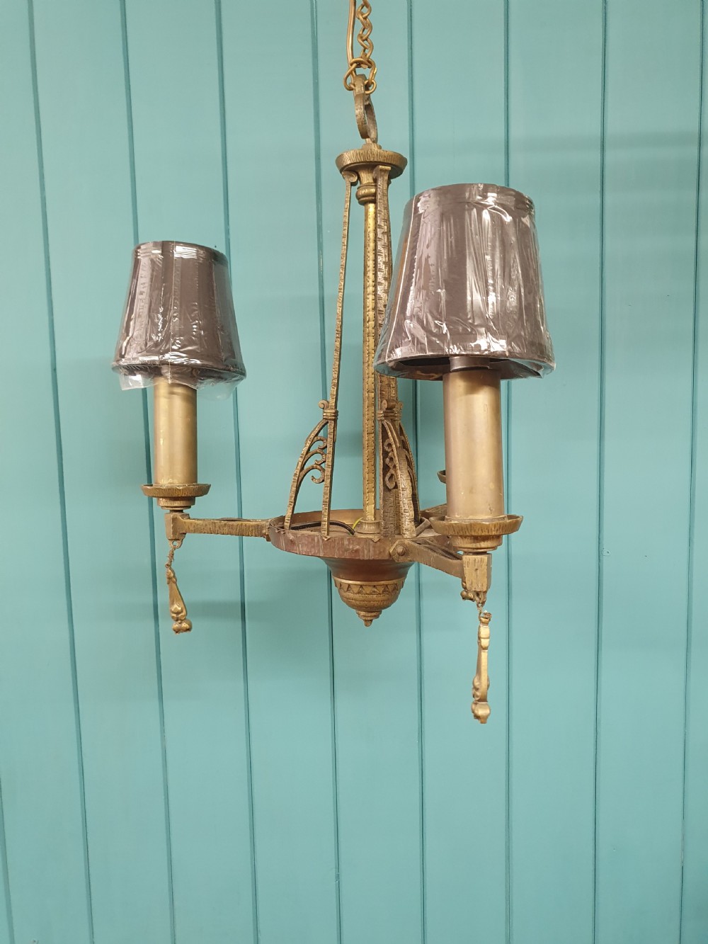 An Art Deco gilt brass three-arm chandelier featuring geometric central columns, candle-style bulb holders, and dangling teardrop accents. View X