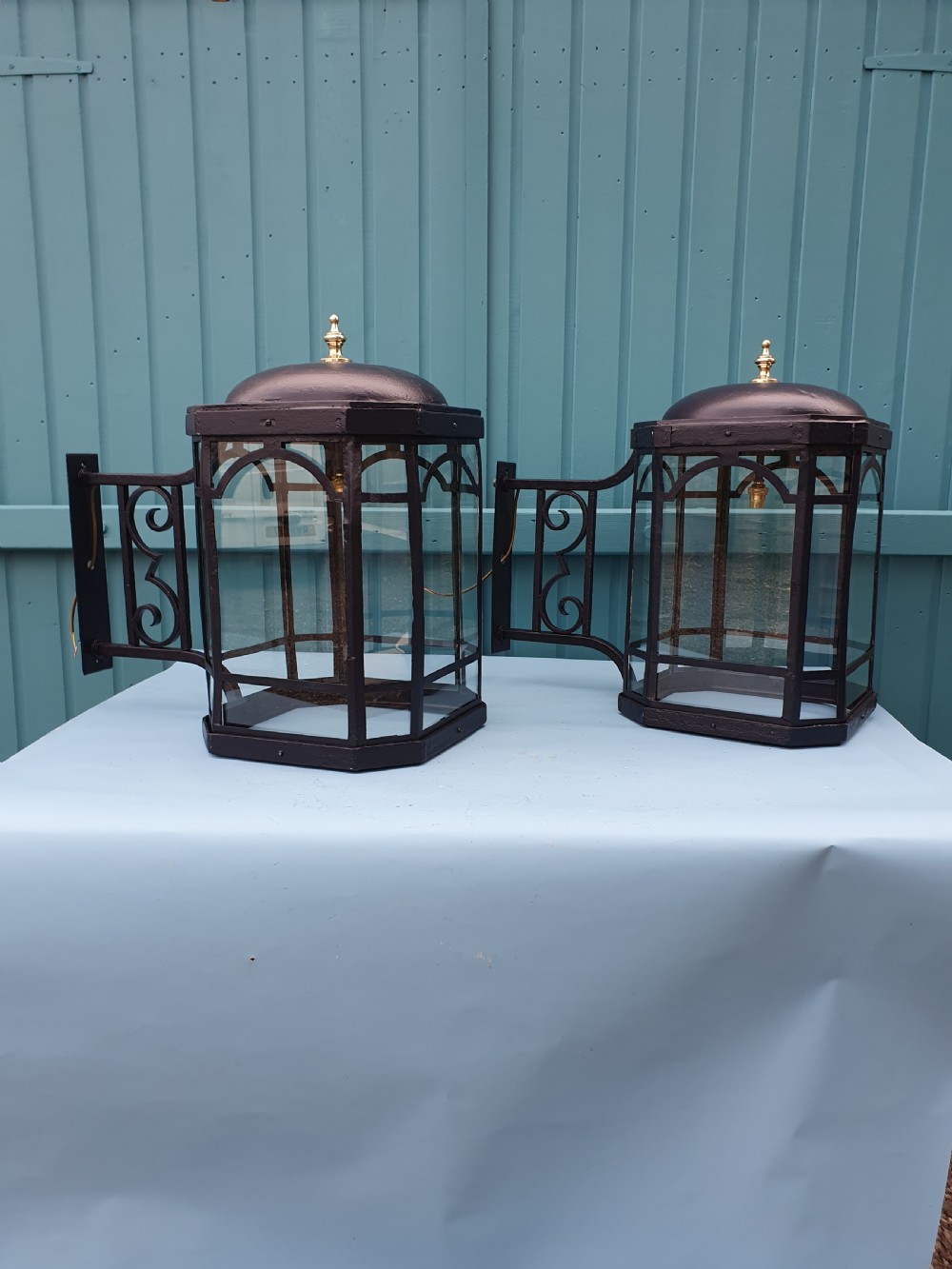 A pair of early 20th-century octagonal black wrought iron wall lanterns featuring clear glass panels, arched tracery, scrollwork brackets, and brass finials.