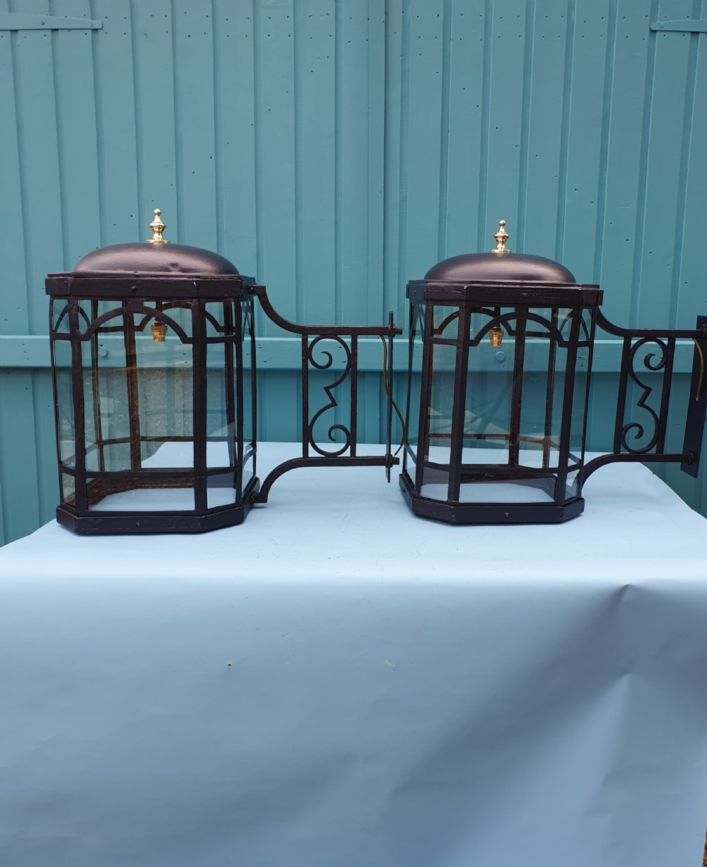 A pair of early 20th-century octagonal black wrought iron wall lanterns featuring clear glass panels, arched tracery, scrollwork brackets, and brass finials. View V