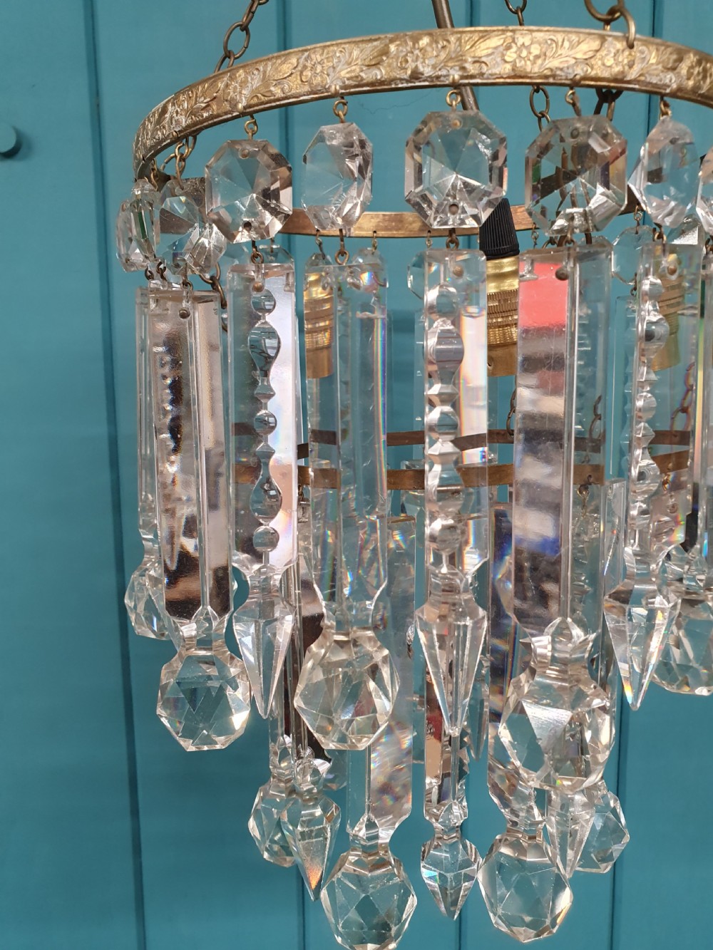 A small early 20th-century ceiling chandelier featuring a decorative brass gallery with tiered, clear glass prism drops hanging from thin metal chains. View VII