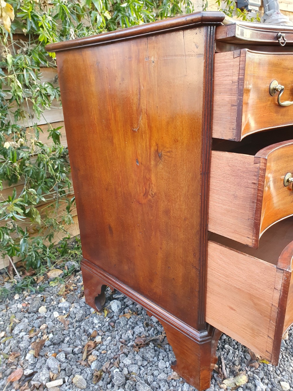 A Georgian mahogany serpentine chest of drawers featuring a curved three-drawer front, brass swan-neck handles, and shaped bracket feet. View VI