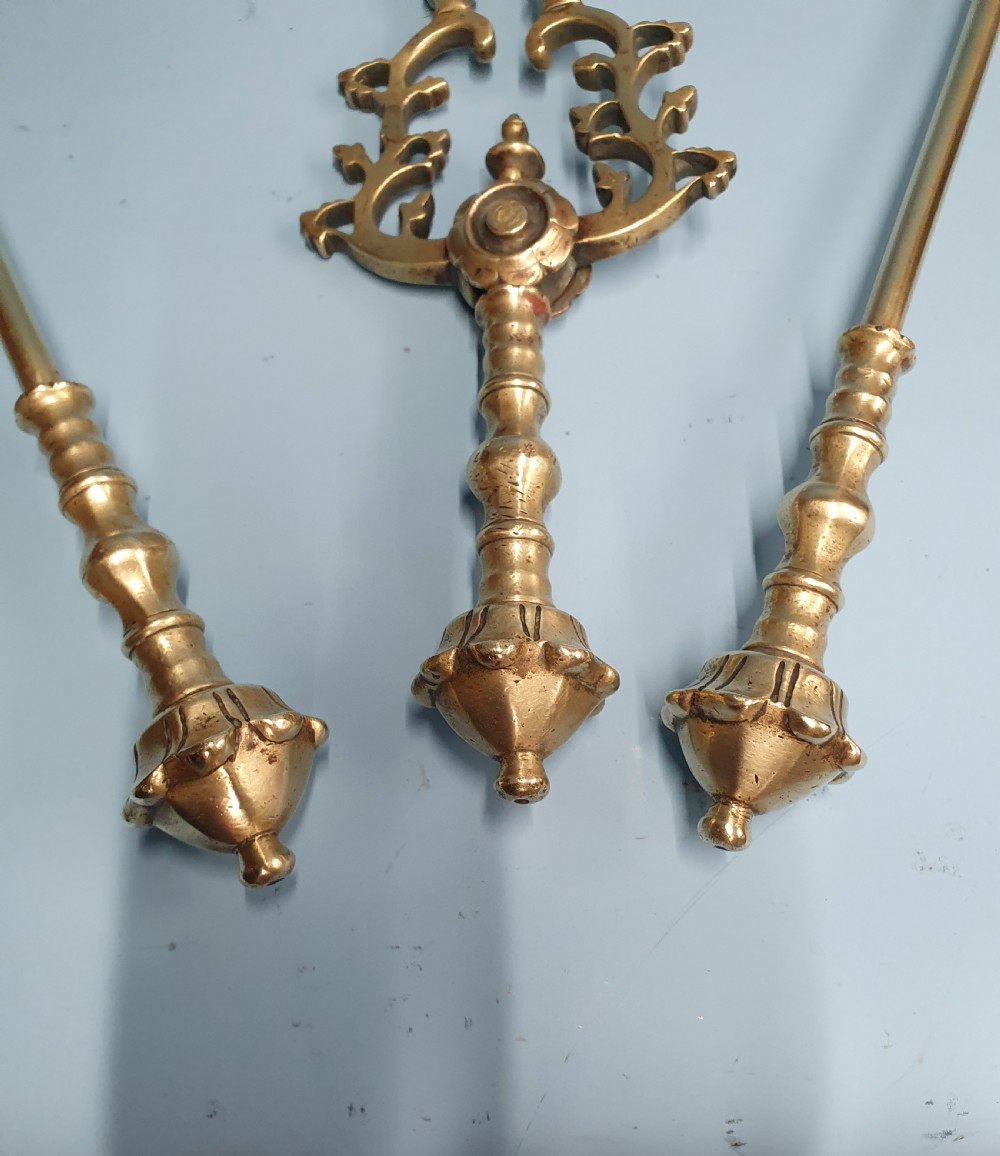 A three-piece set of 19th-century brass fire tools comprising a poker, tongs, and an ornamental shovel with a decorative pierced pattern. View II