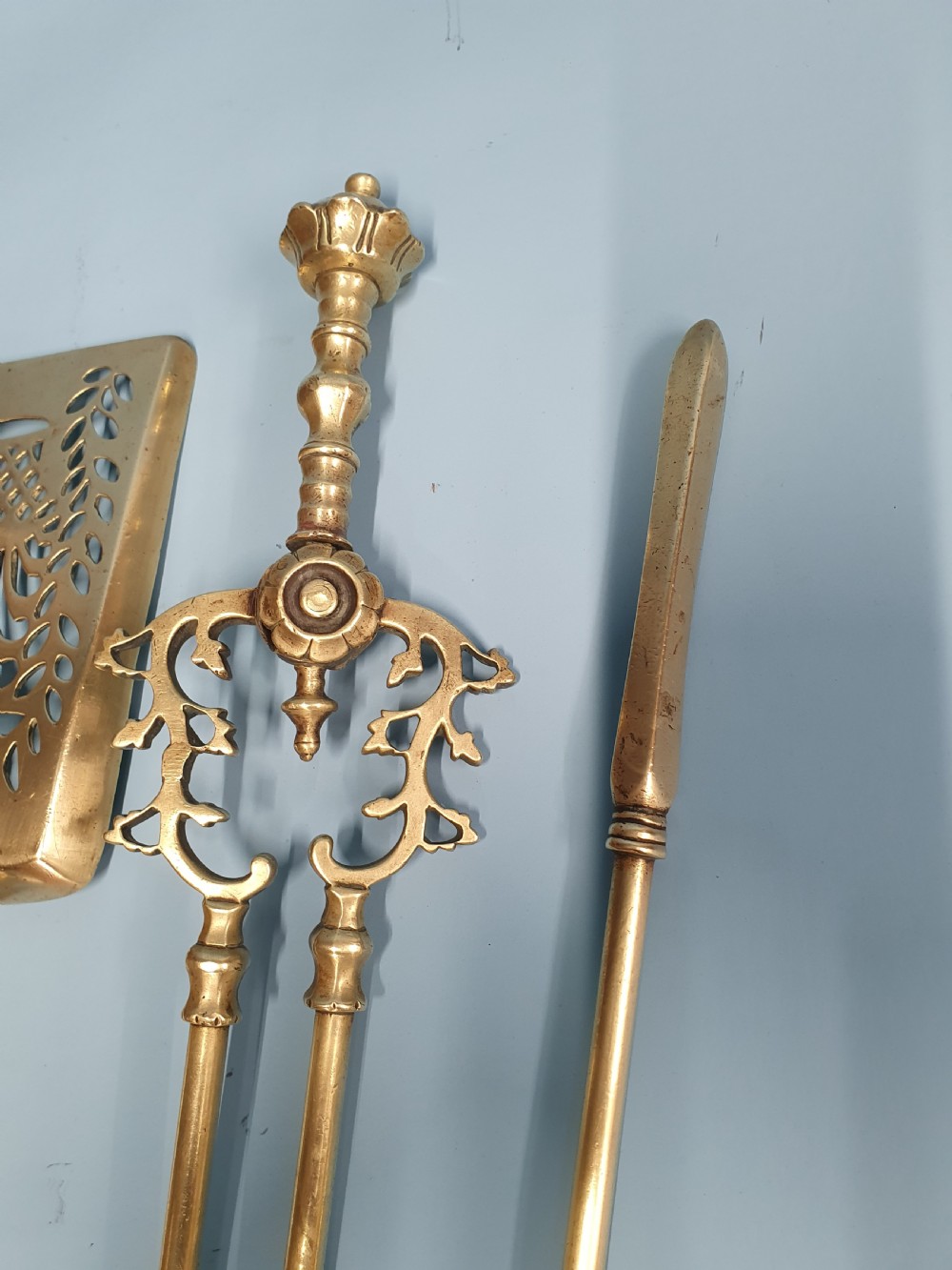A three-piece set of 19th-century brass fire tools comprising a poker, tongs, and an ornamental shovel with a decorative pierced pattern. View IV