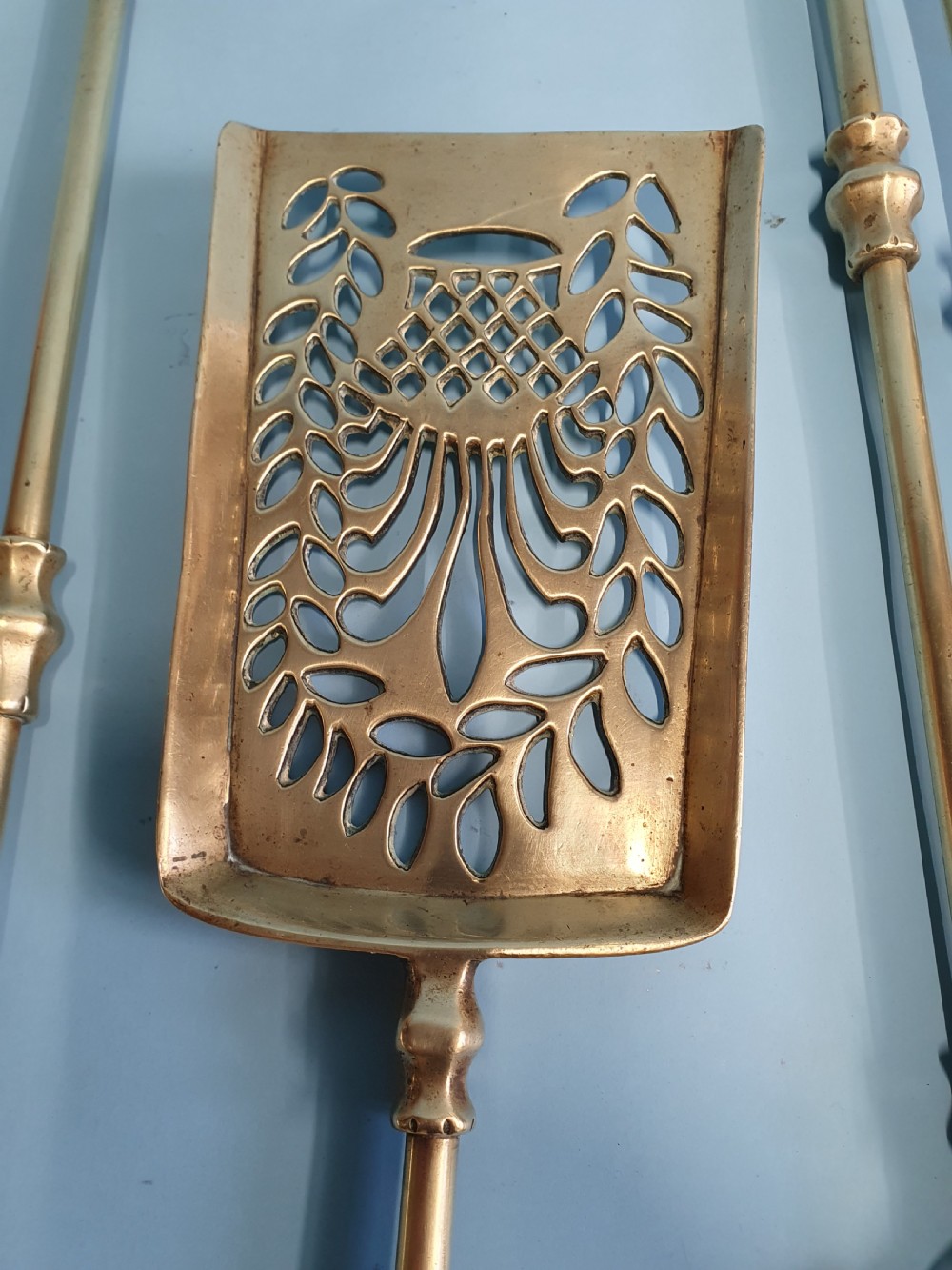 A three-piece set of 19th-century brass fire tools comprising a poker, tongs, and an ornamental shovel with a decorative pierced pattern. View VI