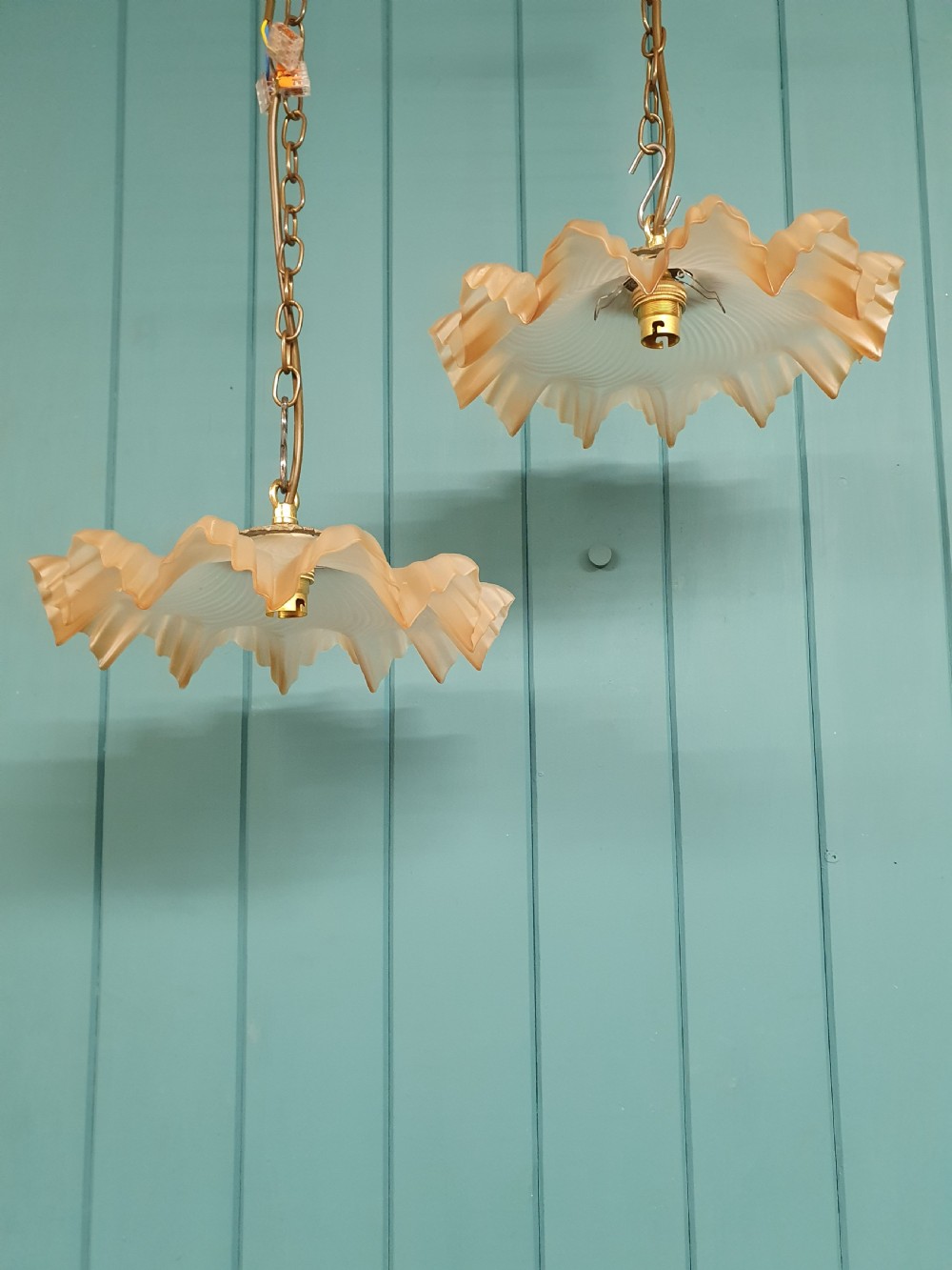 A pair of French pendant lights featuring peach frosted glass shades with ruffled fluted rims suspended from brass chains.