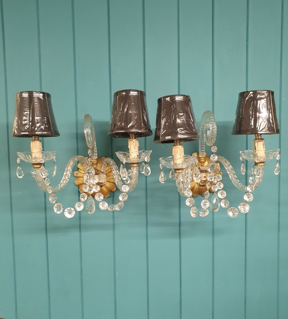 A pair of early 20th-century French double-arm glass wall sconces with crystal droplets, gilt brass backplates, and black lampshades.