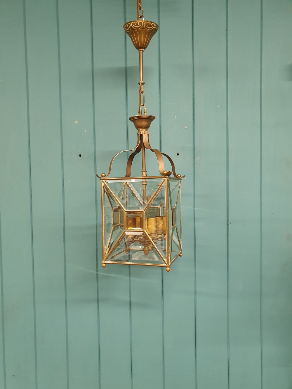 A square brass Arts and Crafts hall lantern featuring geometric bevelled glass panels suspended from a brass chain.