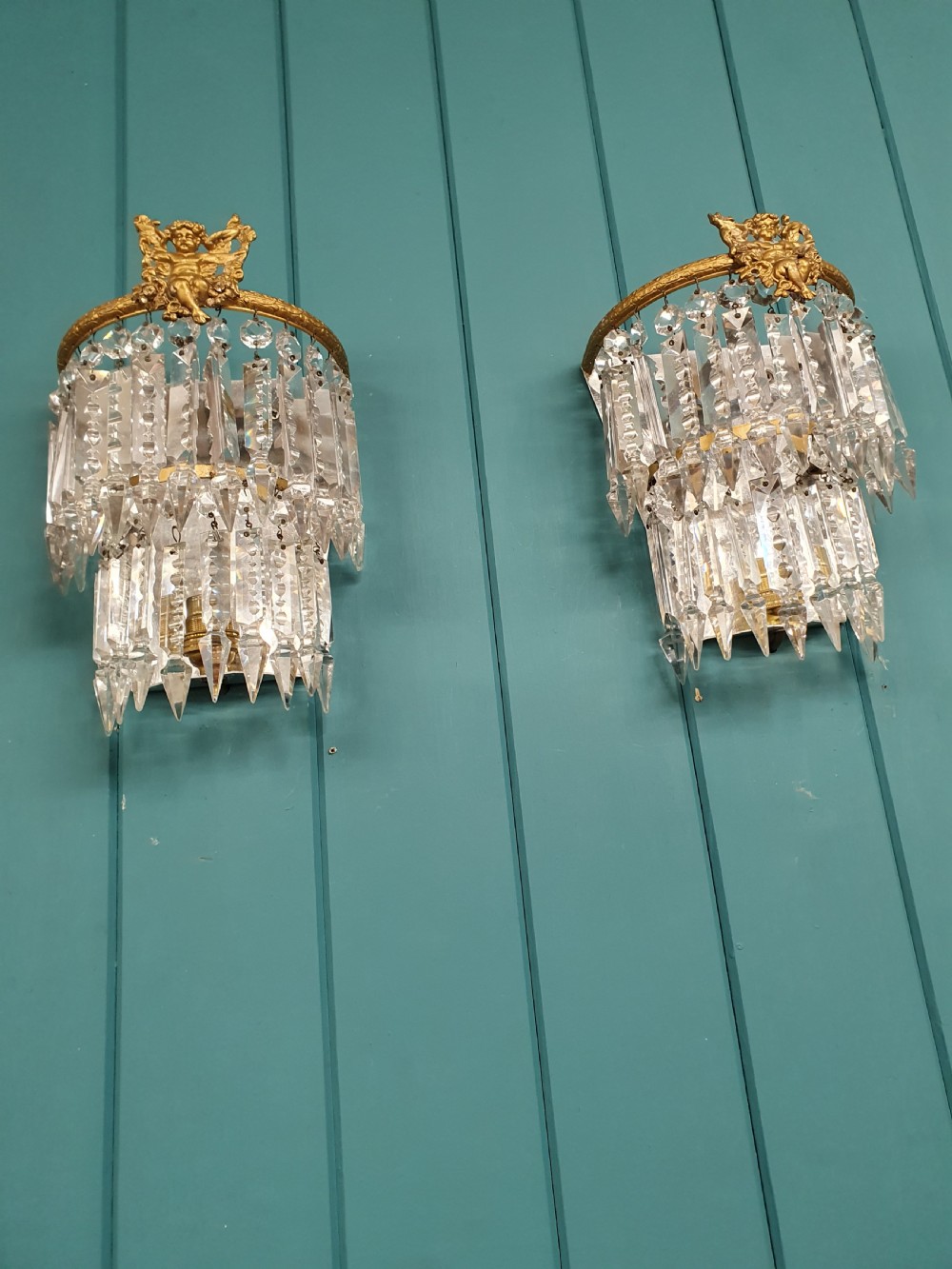 A pair of early 20th-century gilded brass wall sconces featuring top cherub figures and tiered hanging crystal prisms. View V