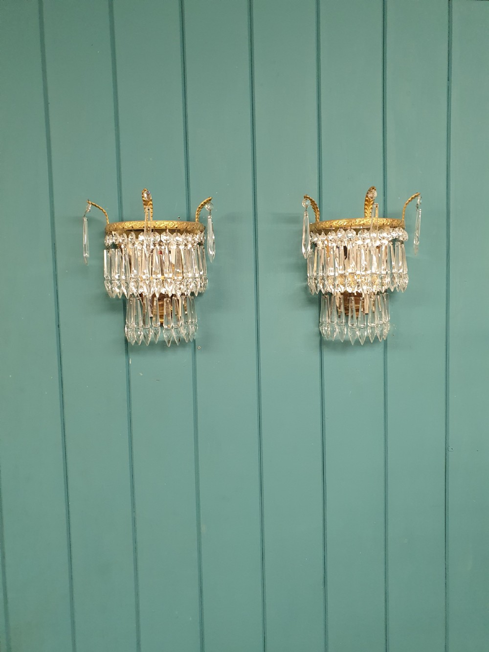 A pair of antique gilt brass wall lights featuring tiered crystal prisms, decorative top spikes, and mirrored backings.