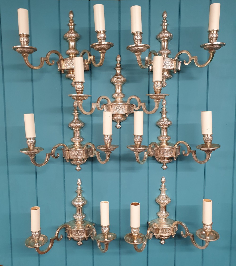 Set of seven silver-plated twin-branch wall lights featuring relief-carved backplates, urn finials, twin scroll arms, and faux candle sleeves.