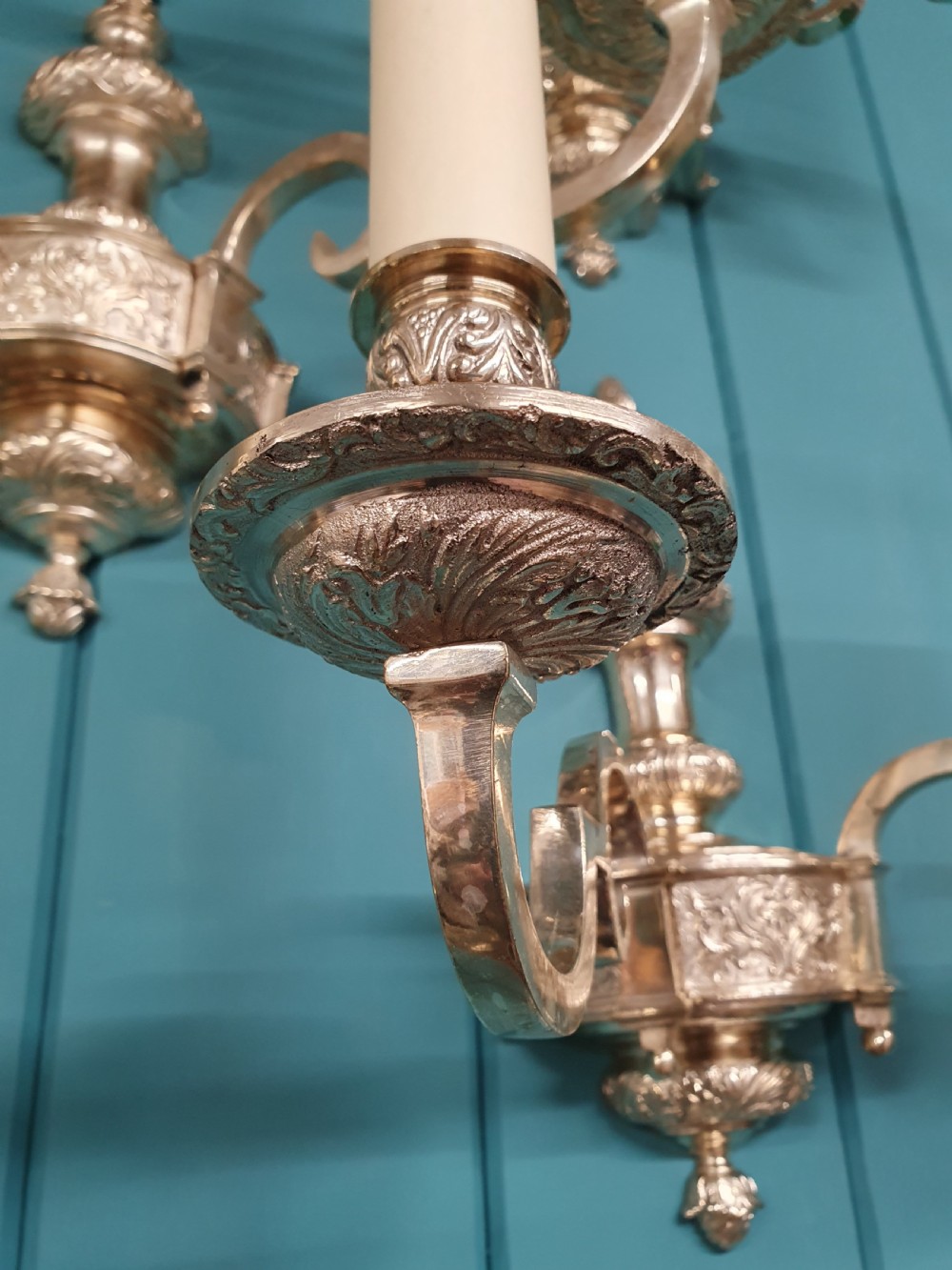 Set of seven silver-plated twin-branch wall lights featuring relief-carved backplates, urn finials, twin scroll arms, and faux candle sleeves. View VI