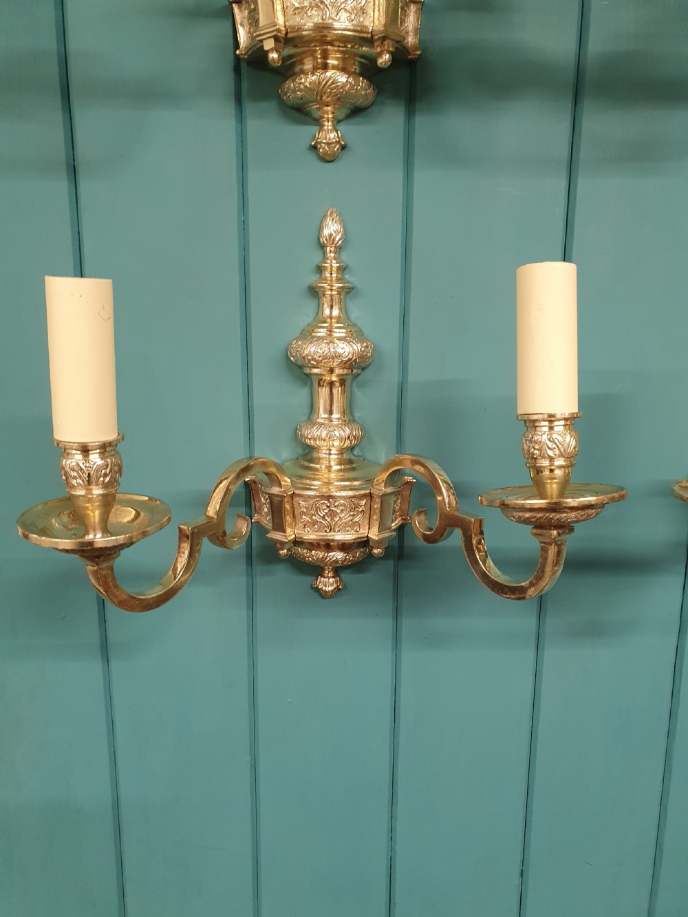 Set of seven silver-plated twin-branch wall lights featuring relief-carved backplates, urn finials, twin scroll arms, and faux candle sleeves. View IX