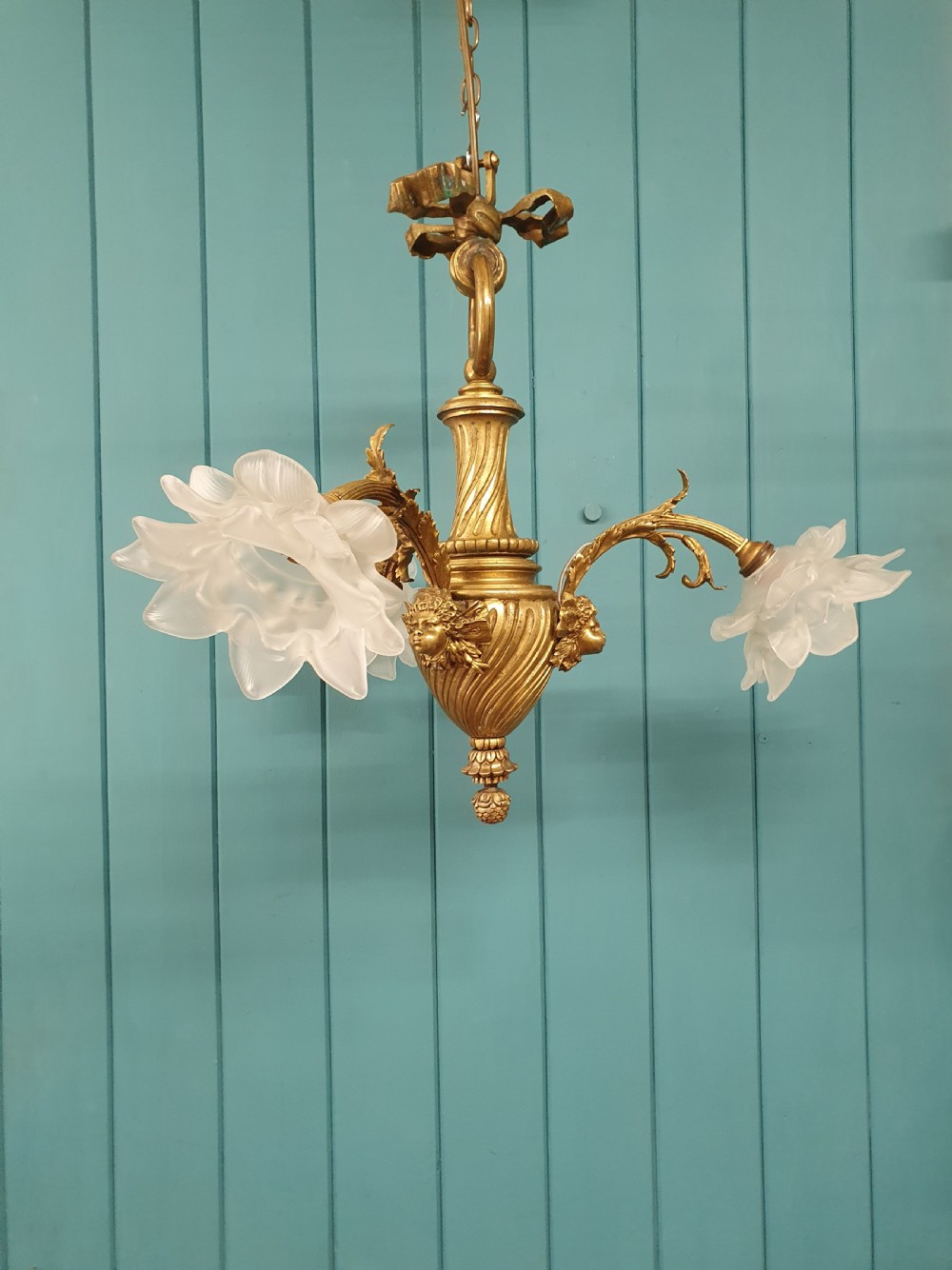A cast brass three-arm chandelier featuring cherub head reliefs on a central fluted body and floral frosted glass shades.