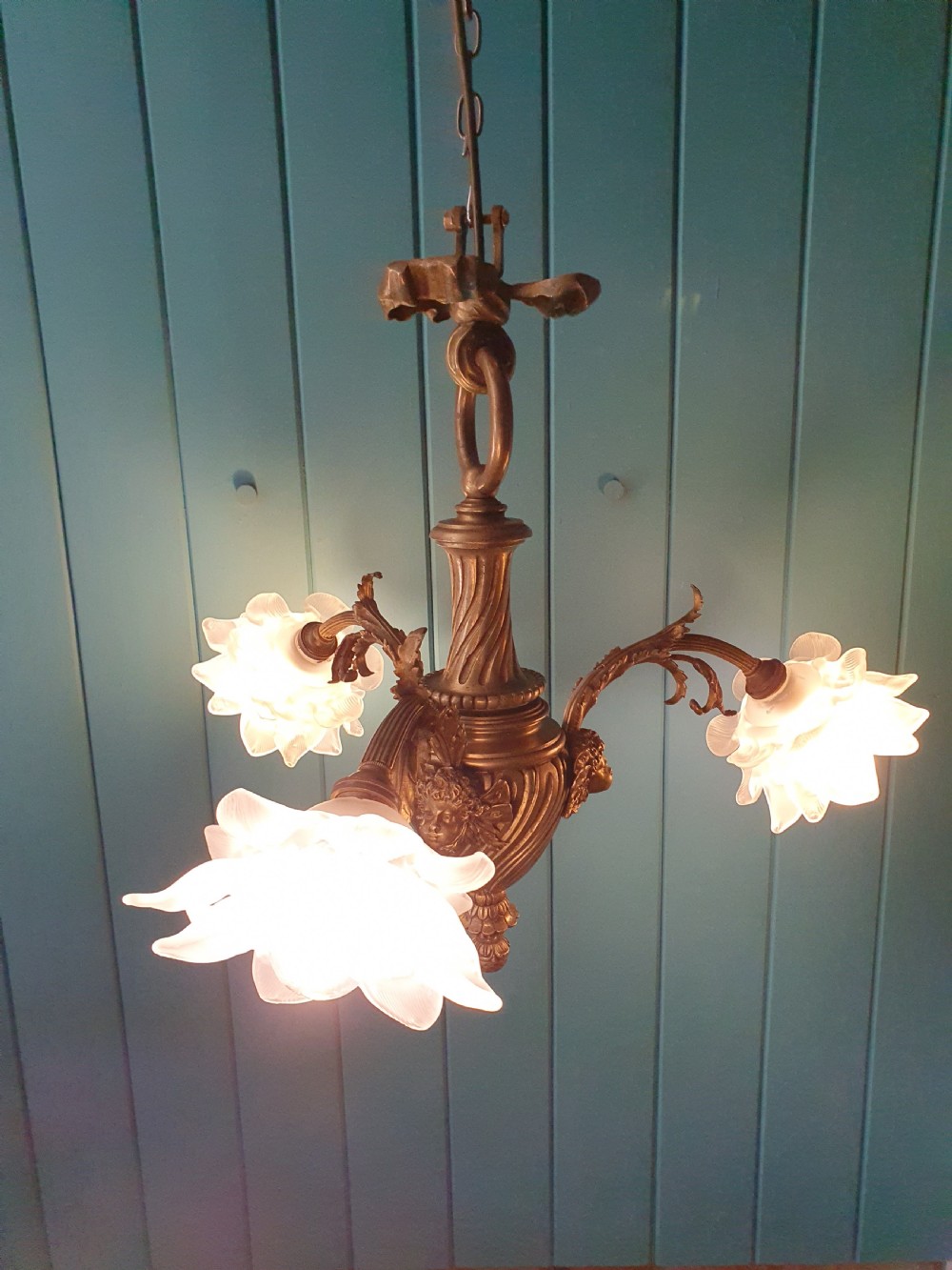 A cast brass three-arm chandelier featuring cherub head reliefs on a central fluted body and floral frosted glass shades. View VII