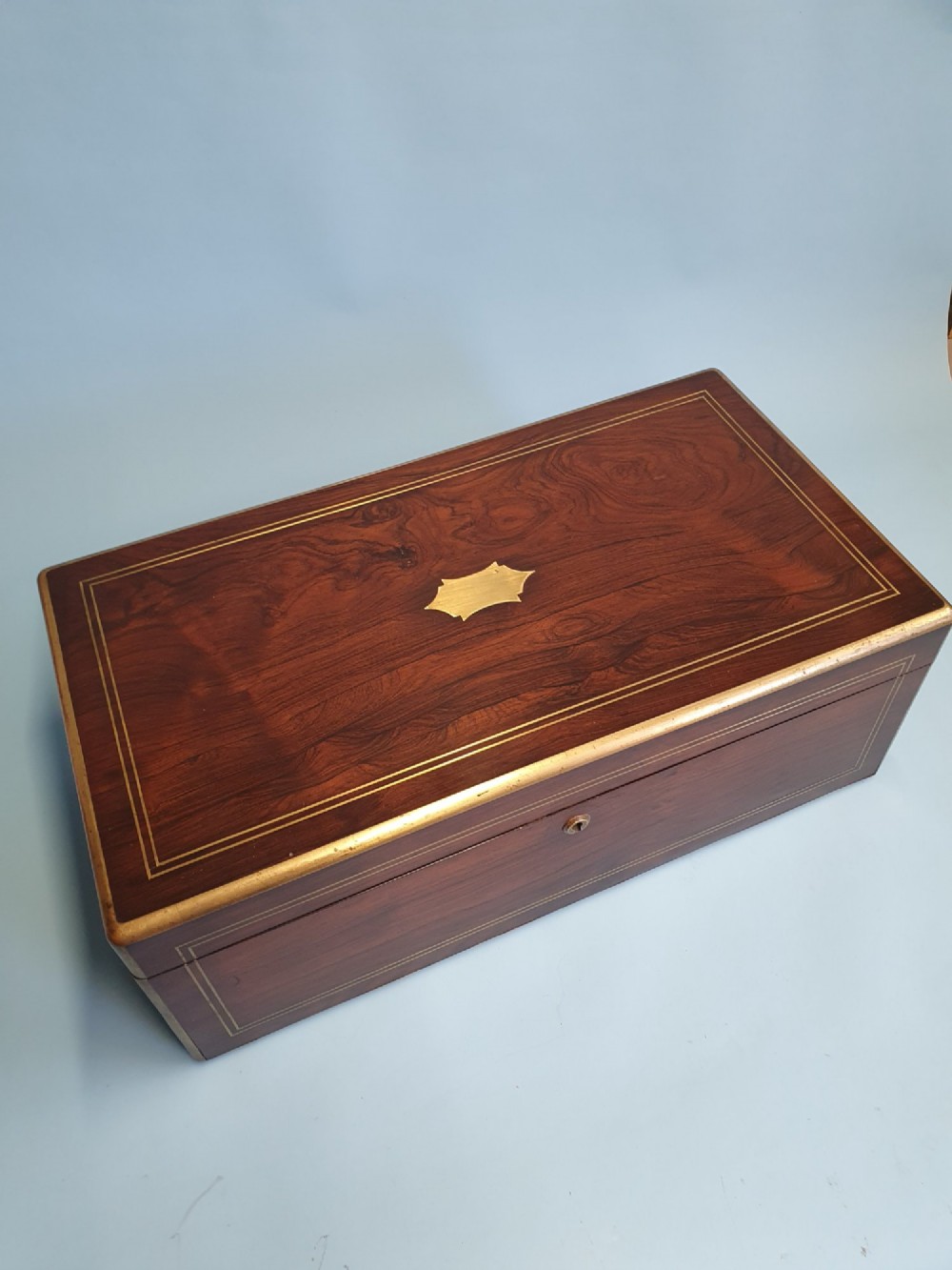 A rectangular dark rosewood writing slope featuring brass inlay stringing, brass corner brackets, a keyhole, and a central brass cartouche. View V