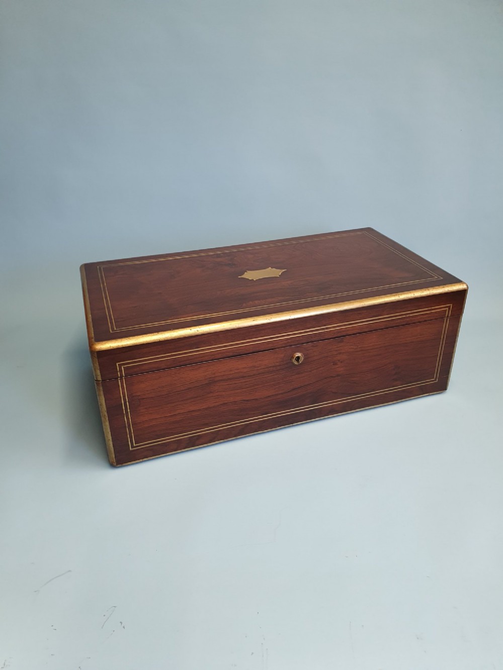 A rectangular dark rosewood writing slope featuring brass inlay stringing, brass corner brackets, a keyhole, and a central brass cartouche.