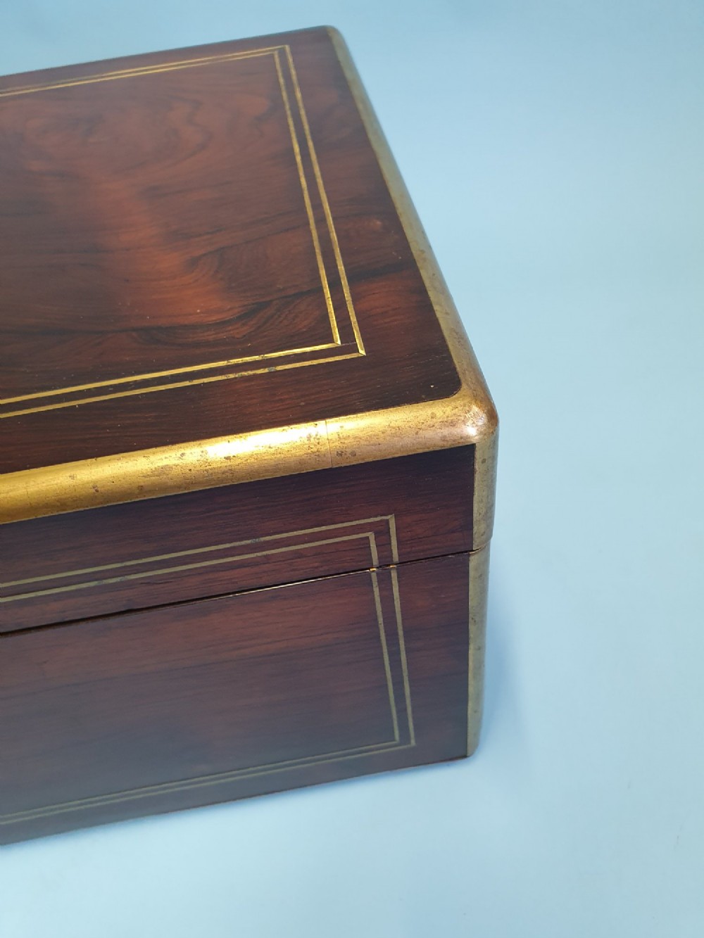 A rectangular dark rosewood writing slope featuring brass inlay stringing, brass corner brackets, a keyhole, and a central brass cartouche. View X