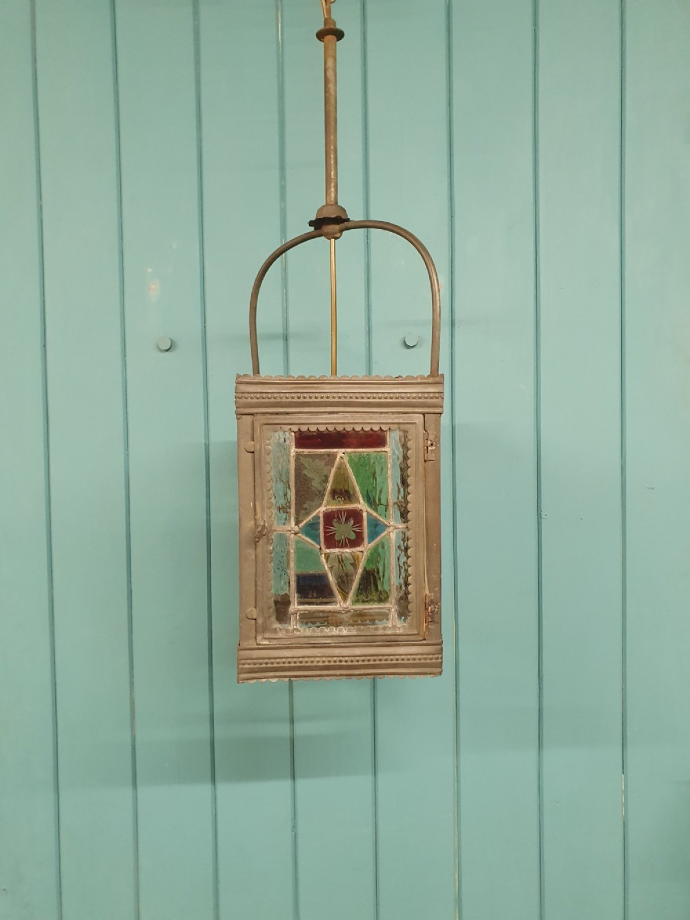 A Victorian hall lantern featuring a rectangular metal frame, an arched suspension handle, and leaded stained glass panels in green, red, and blue geometric patterns.