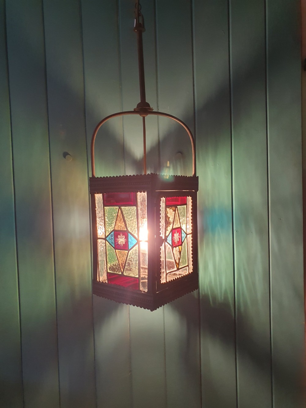 A Victorian hall lantern featuring a rectangular metal frame, an arched suspension handle, and leaded stained glass panels in green, red, and blue geometric patterns. View V
