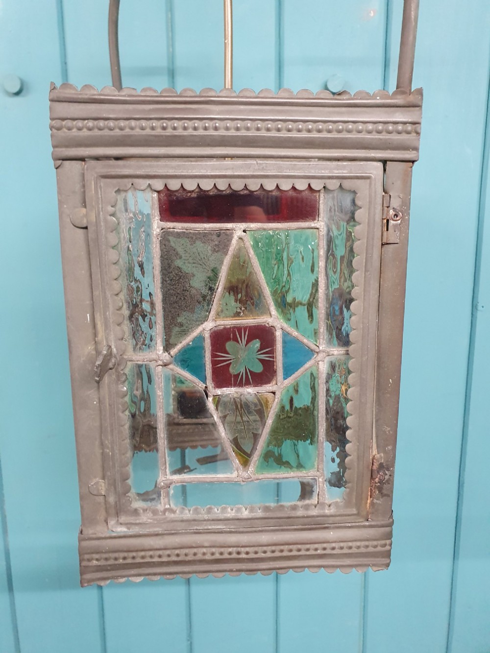 A Victorian hall lantern featuring a rectangular metal frame, an arched suspension handle, and leaded stained glass panels in green, red, and blue geometric patterns. View VI