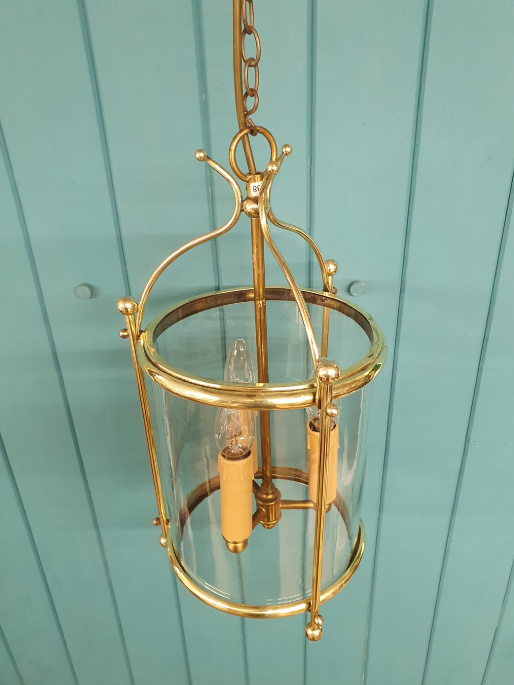 A brass cylindrical hanging hall lantern with clear glass panels, three central candle sleeves, and a suspended brass chain. View III