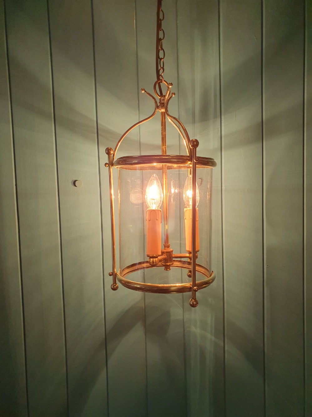 A brass cylindrical hanging hall lantern with clear glass panels, three central candle sleeves, and a suspended brass chain. View V