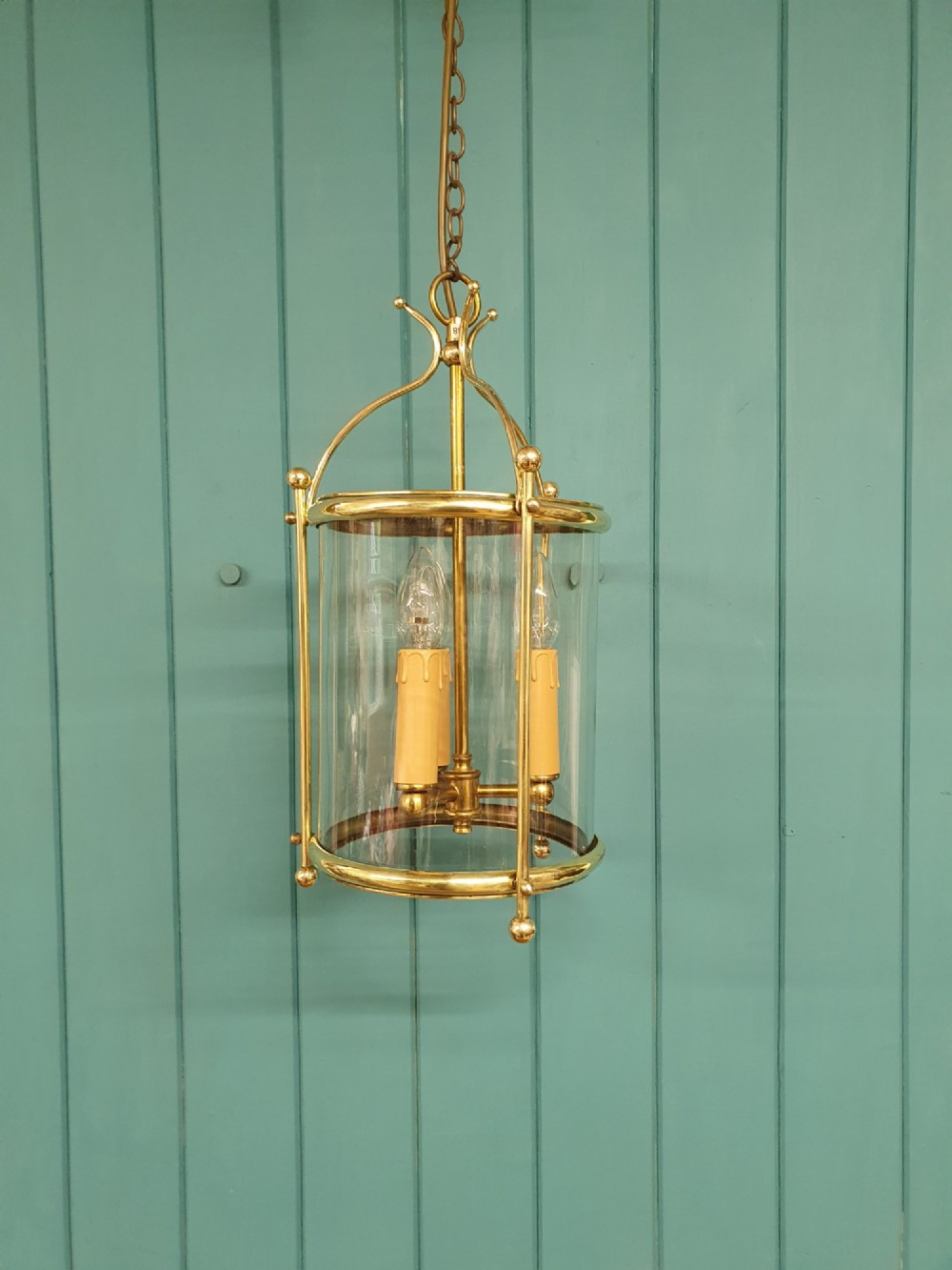 A brass cylindrical hanging hall lantern with clear glass panels, three central candle sleeves, and a suspended brass chain.