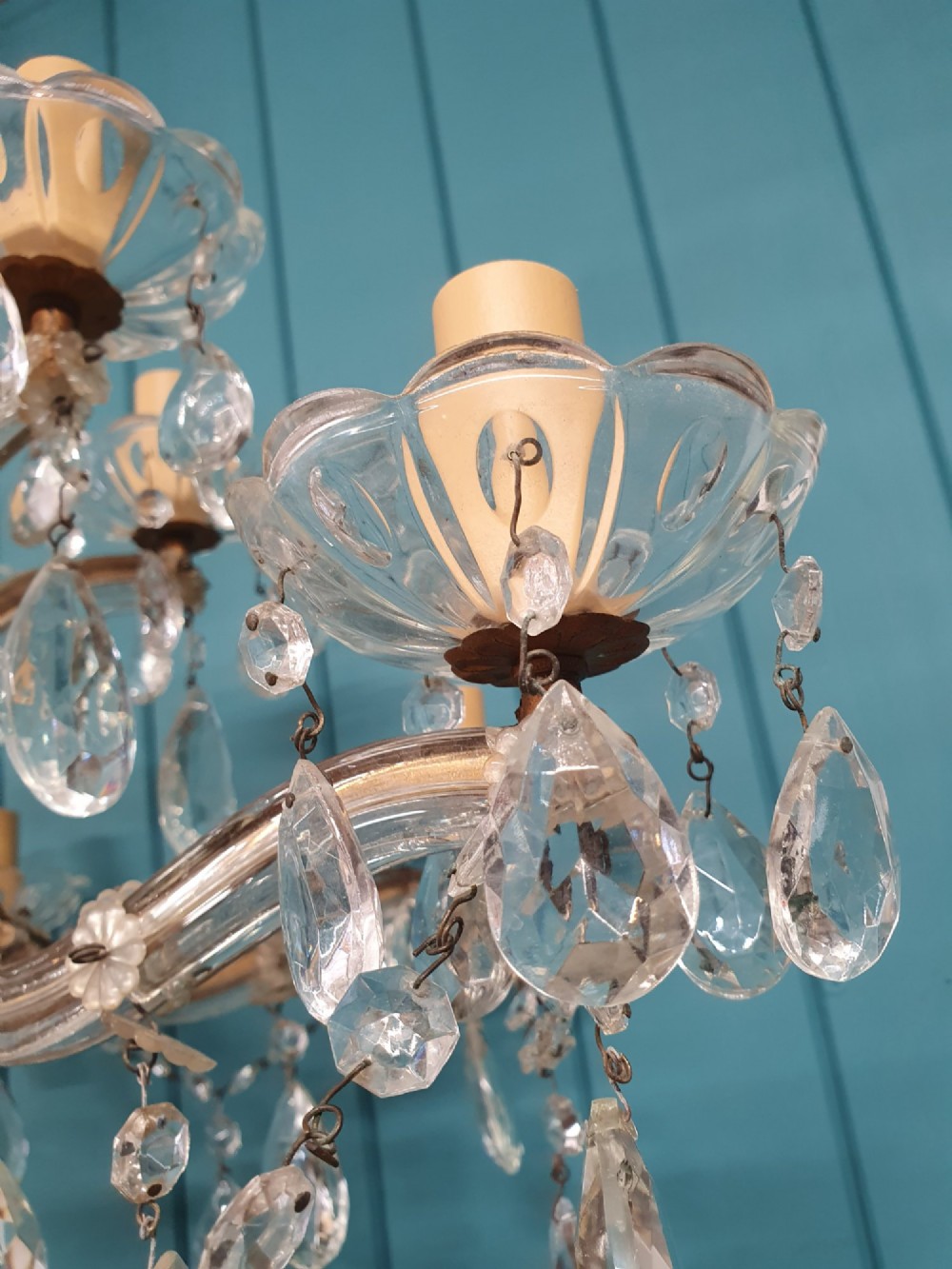 A multi-tiered antique chandelier with twelve curved glass arms, cream candle-style sleeves, and hanging faceted cut crystal teardrop drops against a blue tongue-and-groove background. View II