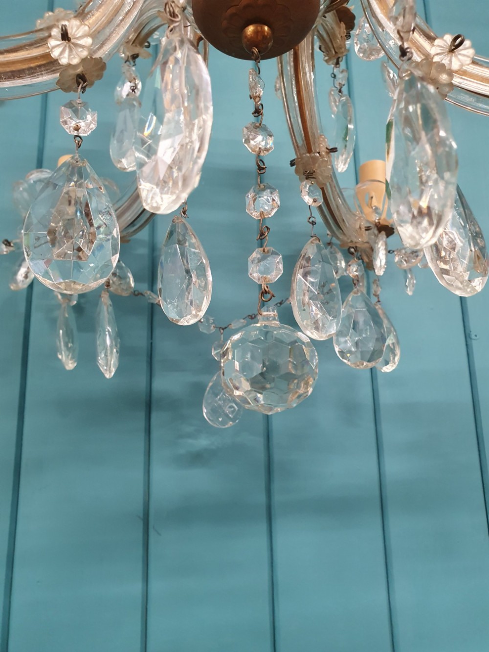 A multi-tiered antique chandelier with twelve curved glass arms, cream candle-style sleeves, and hanging faceted cut crystal teardrop drops against a blue tongue-and-groove background. View III