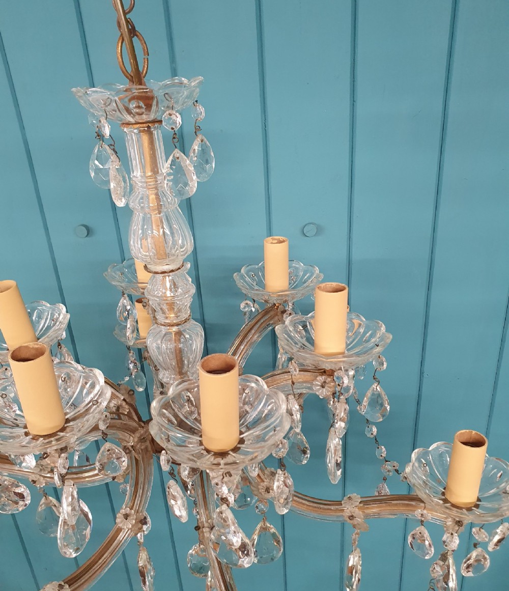 A multi-tiered antique chandelier with twelve curved glass arms, cream candle-style sleeves, and hanging faceted cut crystal teardrop drops against a blue tongue-and-groove background. View IV