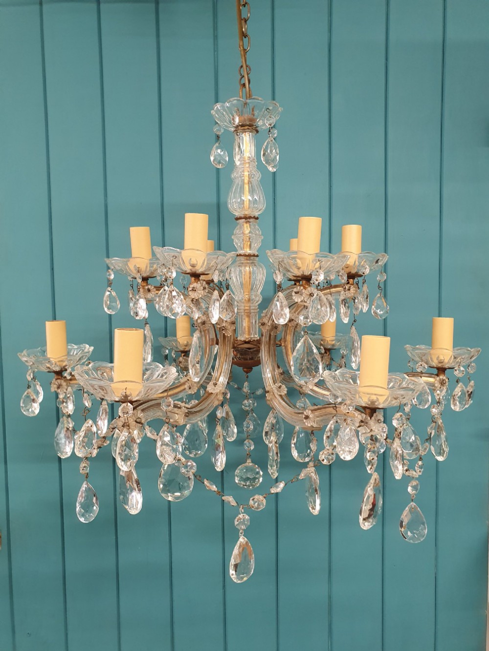 A multi-tiered antique chandelier with twelve curved glass arms, cream candle-style sleeves, and hanging faceted cut crystal teardrop drops against a blue tongue-and-groove background.