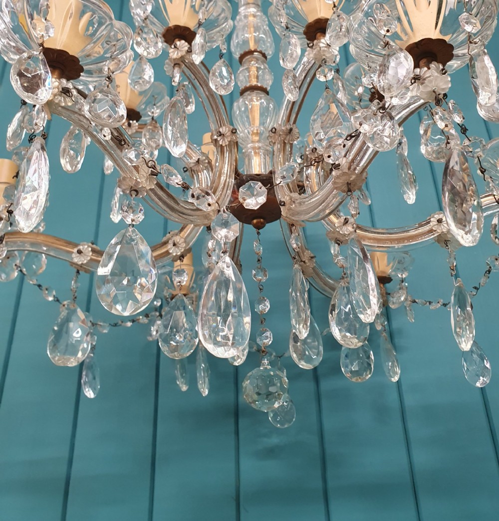 A multi-tiered antique chandelier with twelve curved glass arms, cream candle-style sleeves, and hanging faceted cut crystal teardrop drops against a blue tongue-and-groove background. View V