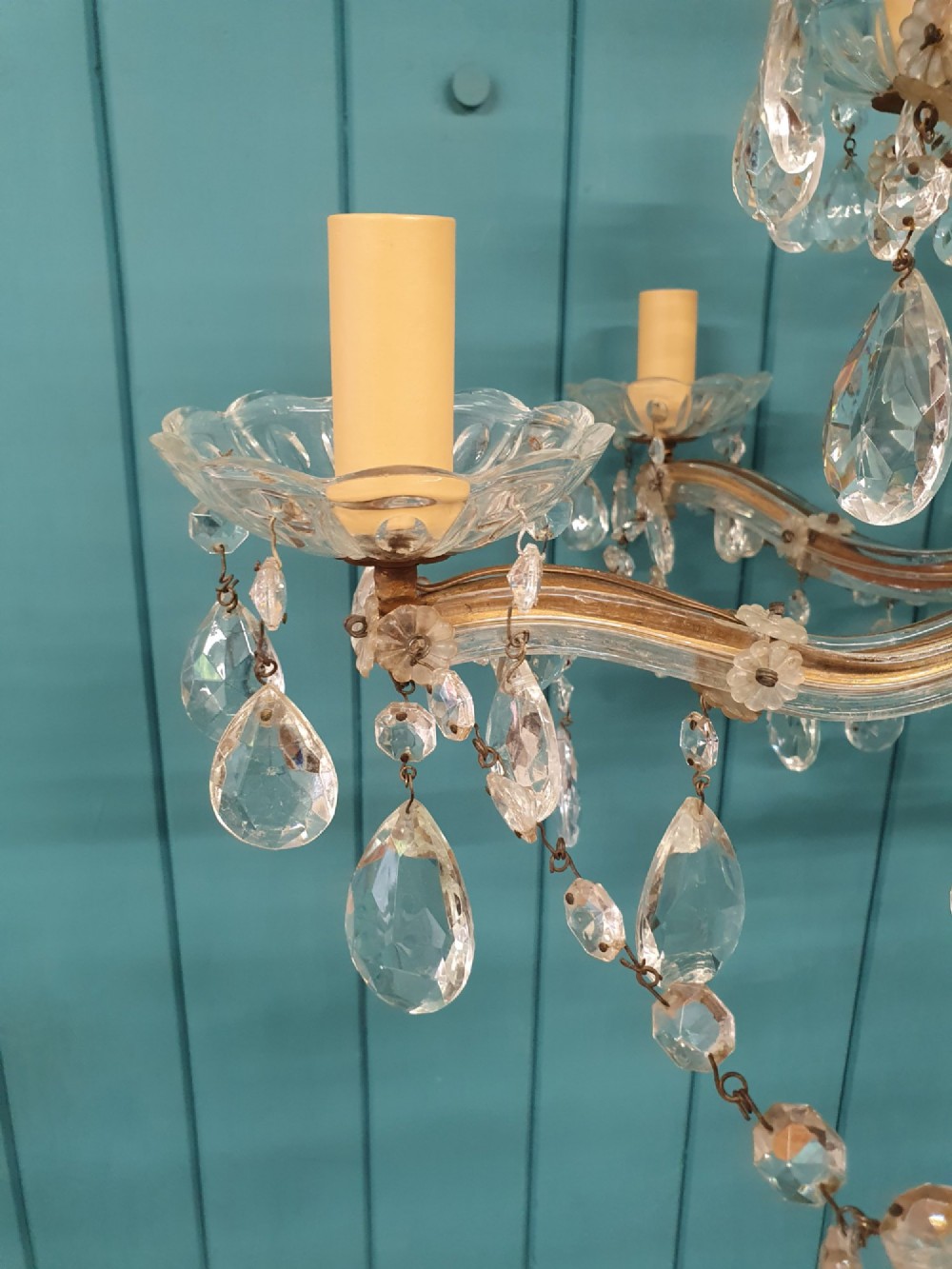 A multi-tiered antique chandelier with twelve curved glass arms, cream candle-style sleeves, and hanging faceted cut crystal teardrop drops against a blue tongue-and-groove background. View VI