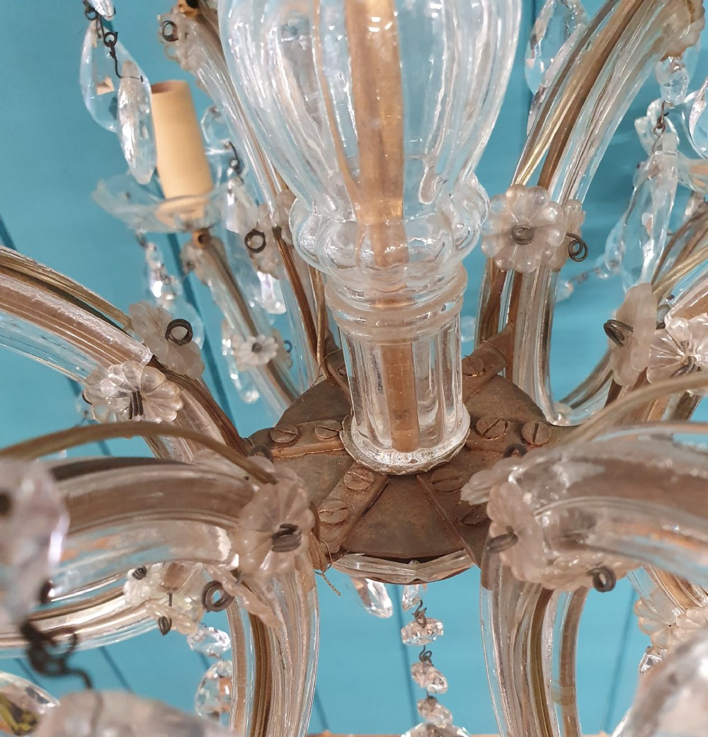 A multi-tiered antique chandelier with twelve curved glass arms, cream candle-style sleeves, and hanging faceted cut crystal teardrop drops against a blue tongue-and-groove background. View VII
