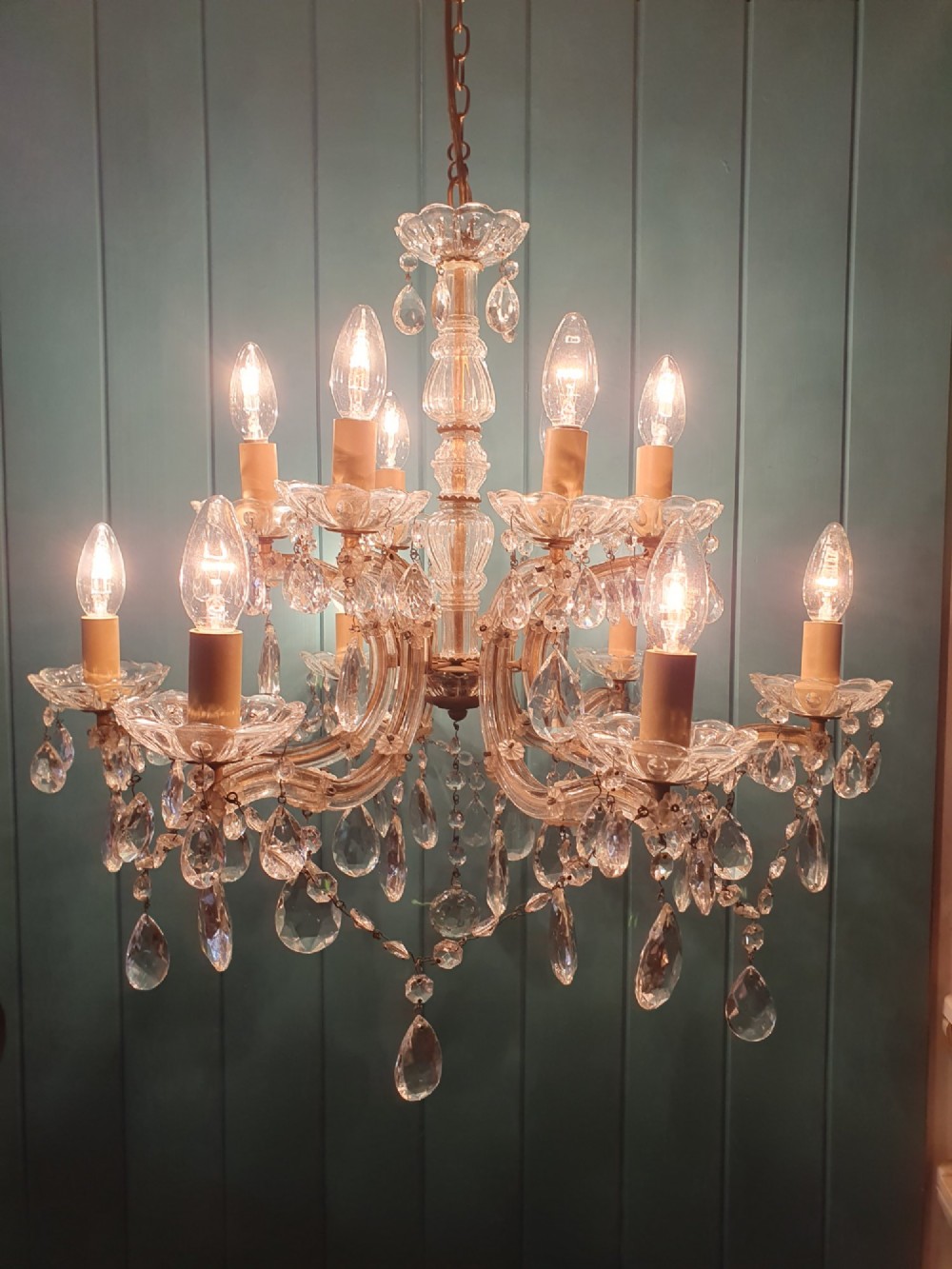 A multi-tiered antique chandelier with twelve curved glass arms, cream candle-style sleeves, and hanging faceted cut crystal teardrop drops against a blue tongue-and-groove background. View VIII
