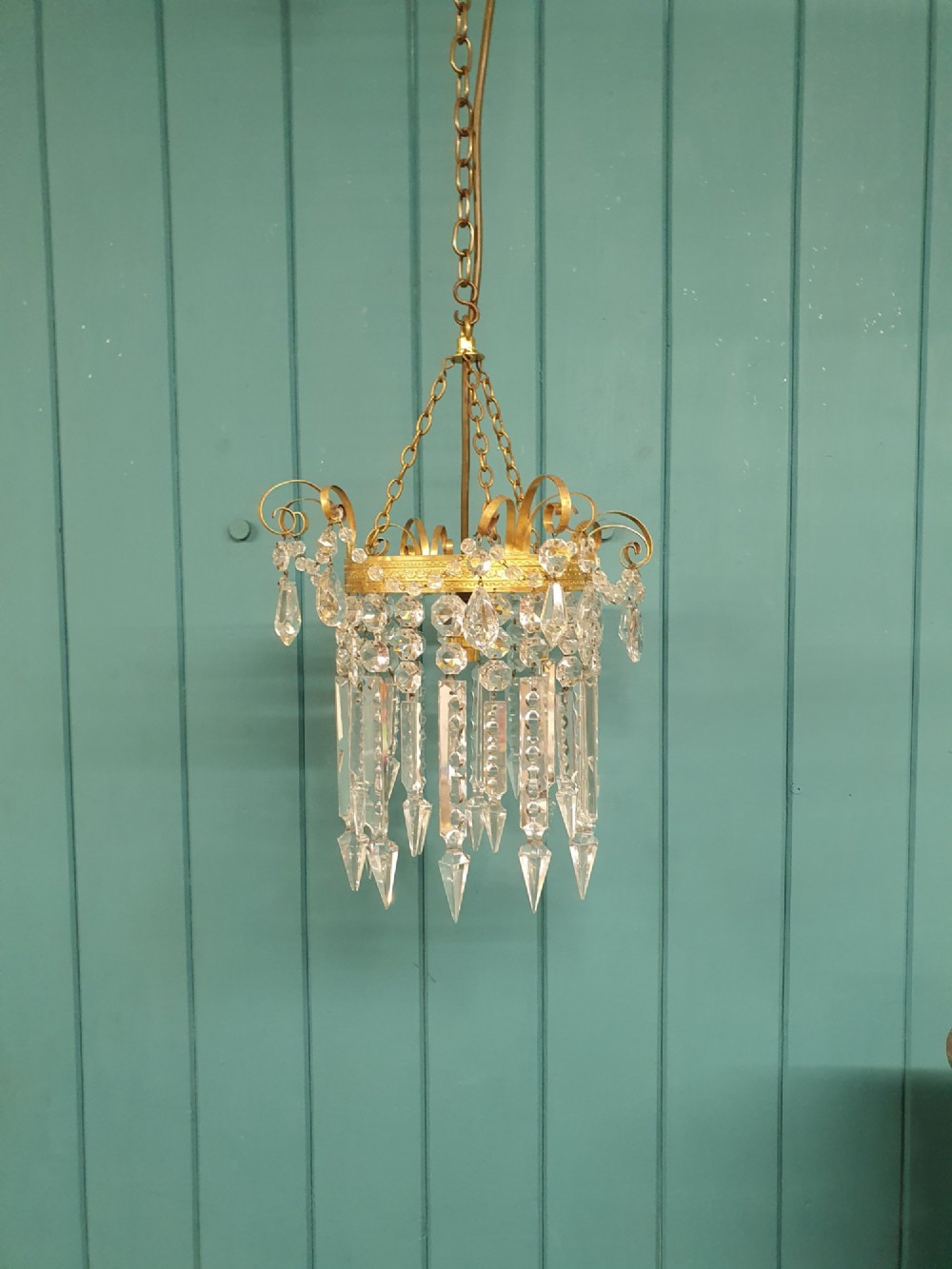 An antique ceiling chandelier featuring a brass metal frame with top scrollwork and tiered cut glass spike prisms hanging down.
