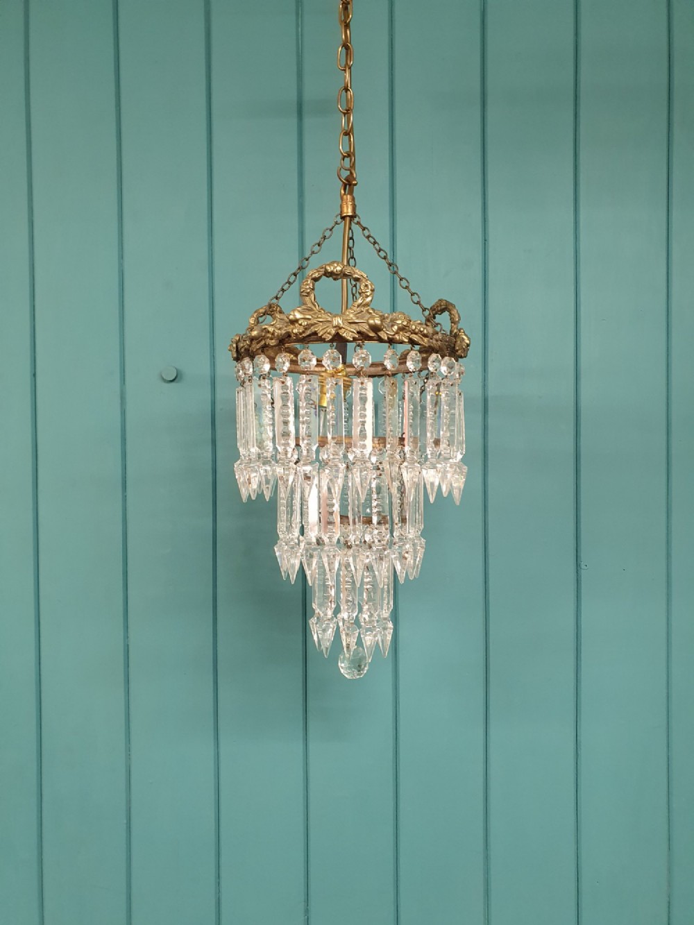 An early 20th-century tiered glass lustre chandelier suspended from a chain, featuring concentric rows of faceted crystal drops below a brass gallery decorated with ribbon motifs.