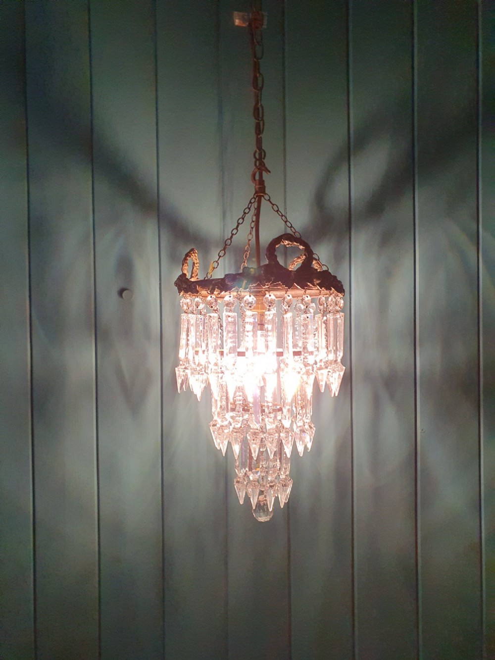 An early 20th-century tiered glass lustre chandelier suspended from a chain, featuring concentric rows of faceted crystal drops below a brass gallery decorated with ribbon motifs. View II