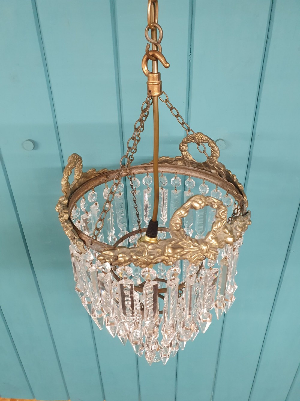 An early 20th-century tiered glass lustre chandelier suspended from a chain, featuring concentric rows of faceted crystal drops below a brass gallery decorated with ribbon motifs. View III