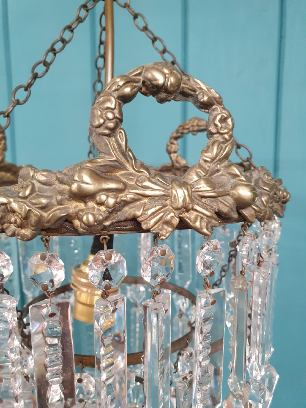 An early 20th-century tiered glass lustre chandelier suspended from a chain, featuring concentric rows of faceted crystal drops below a brass gallery decorated with ribbon motifs. View IV