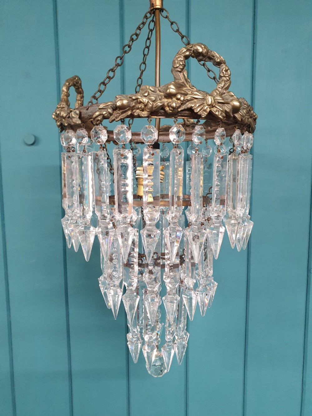 An early 20th-century tiered glass lustre chandelier suspended from a chain, featuring concentric rows of faceted crystal drops below a brass gallery decorated with ribbon motifs. View V