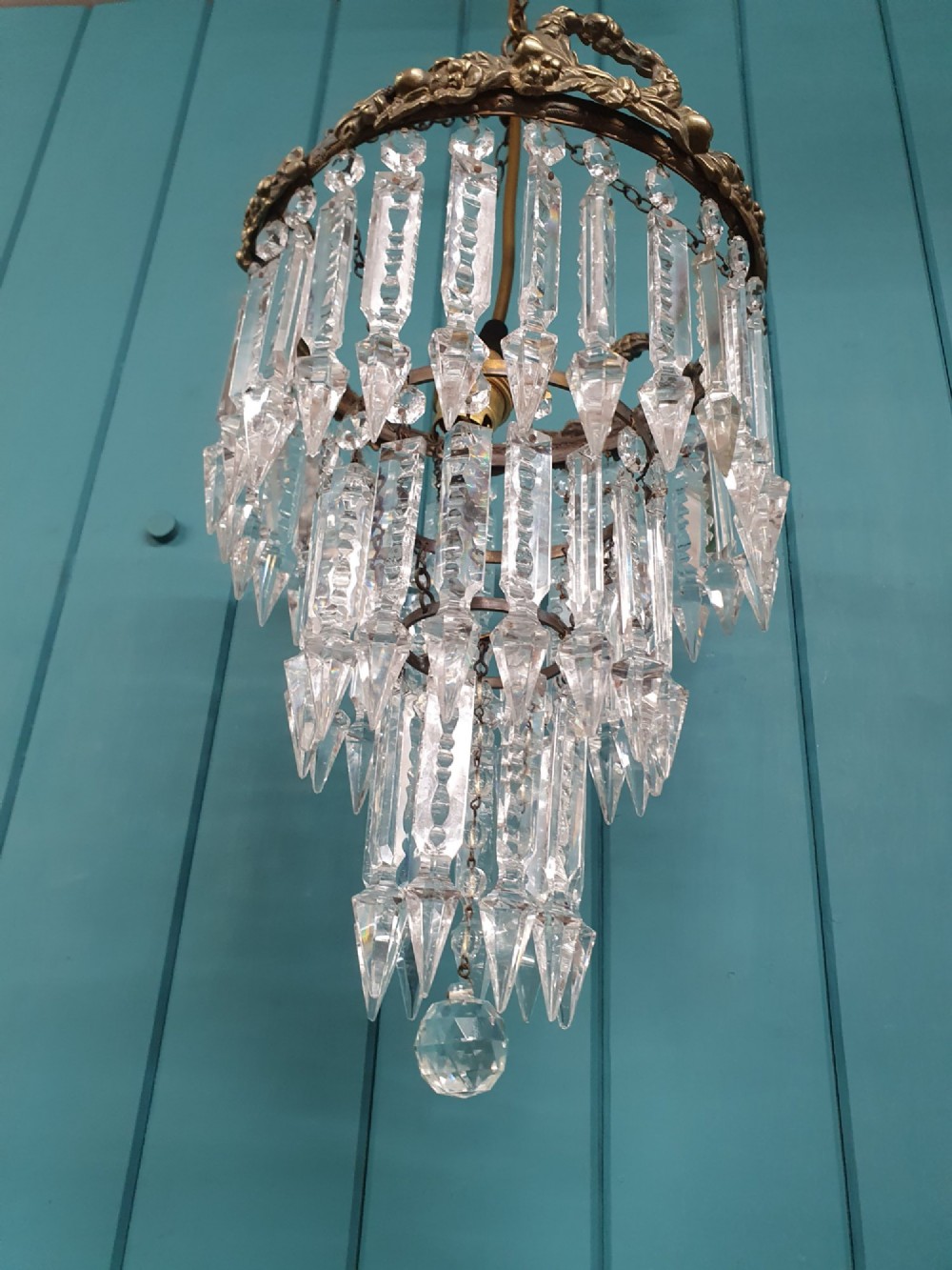 An early 20th-century tiered glass lustre chandelier suspended from a chain, featuring concentric rows of faceted crystal drops below a brass gallery decorated with ribbon motifs. View VI