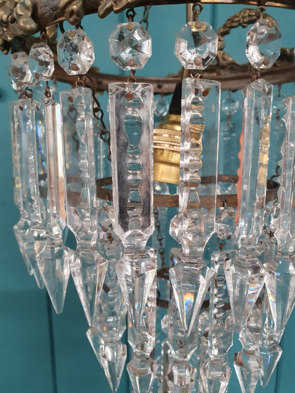 An early 20th-century tiered glass lustre chandelier suspended from a chain, featuring concentric rows of faceted crystal drops below a brass gallery decorated with ribbon motifs. View VII