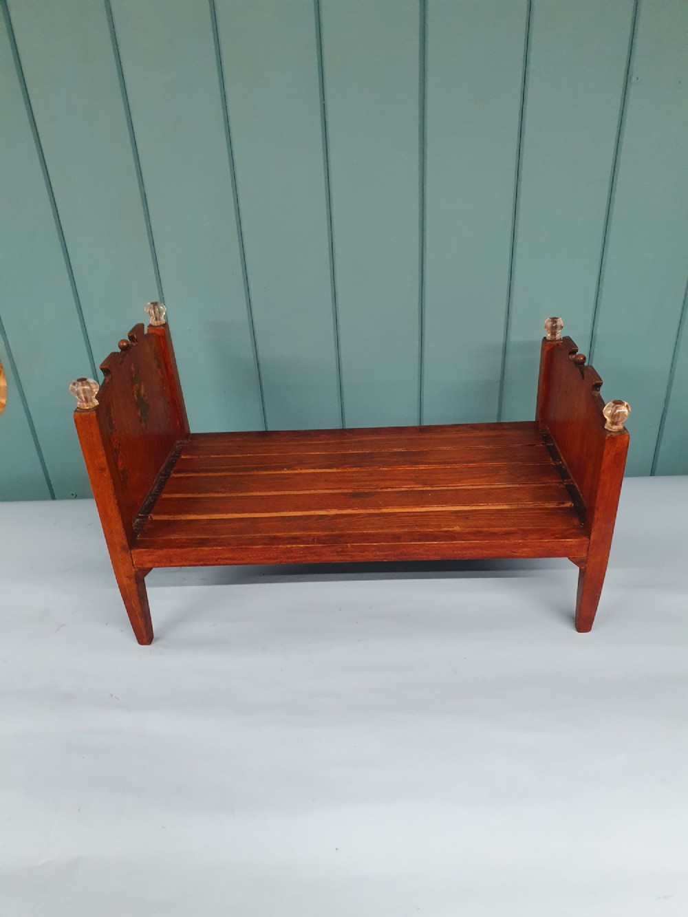A small antique wooden doll bed with hand-painted floral motifs on the headboard and footboard, a slatted frame, and clear glass finials on four corner posts. View II