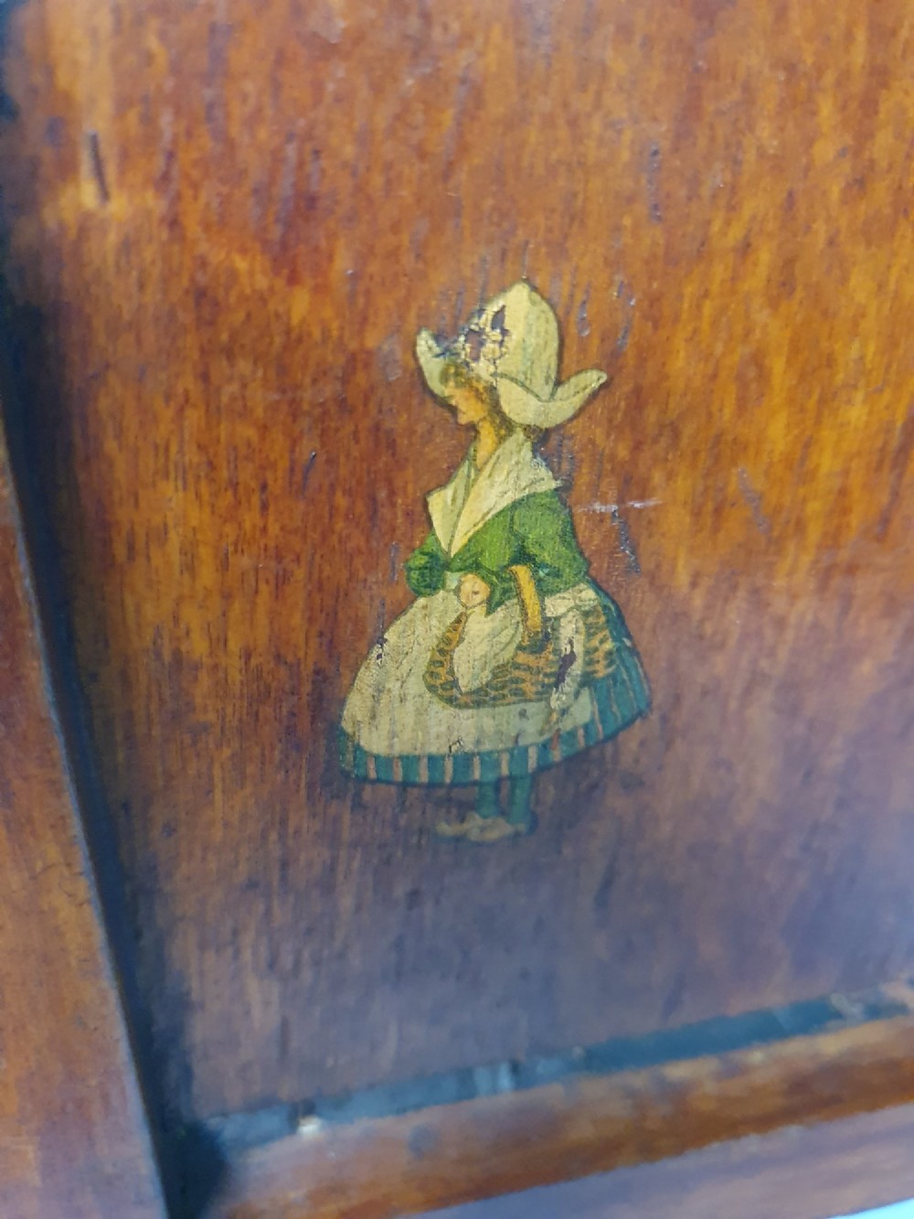 A small antique wooden doll bed with hand-painted floral motifs on the headboard and footboard, a slatted frame, and clear glass finials on four corner posts. View III