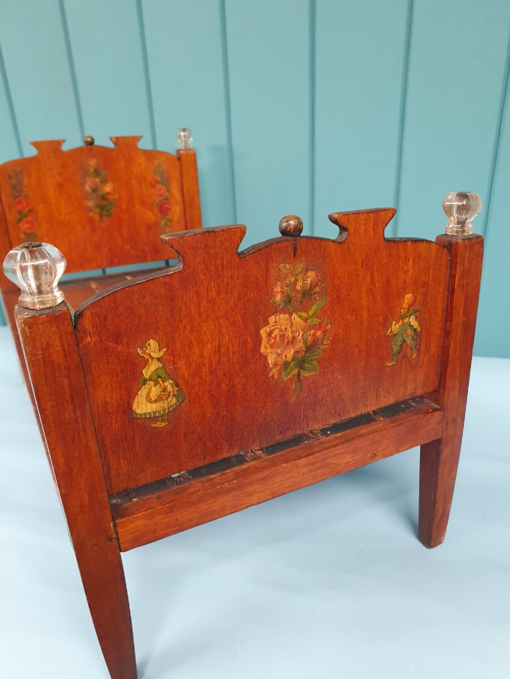 A small antique wooden doll bed with hand-painted floral motifs on the headboard and footboard, a slatted frame, and clear glass finials on four corner posts. View V