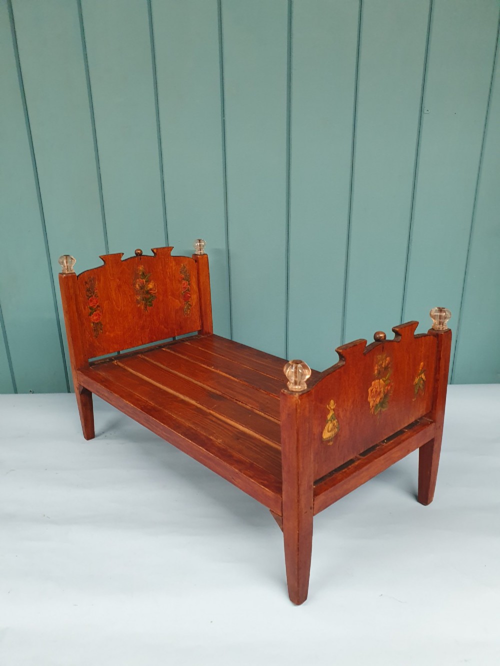 A small antique wooden doll bed with hand-painted floral motifs on the headboard and footboard, a slatted frame, and clear glass finials on four corner posts.